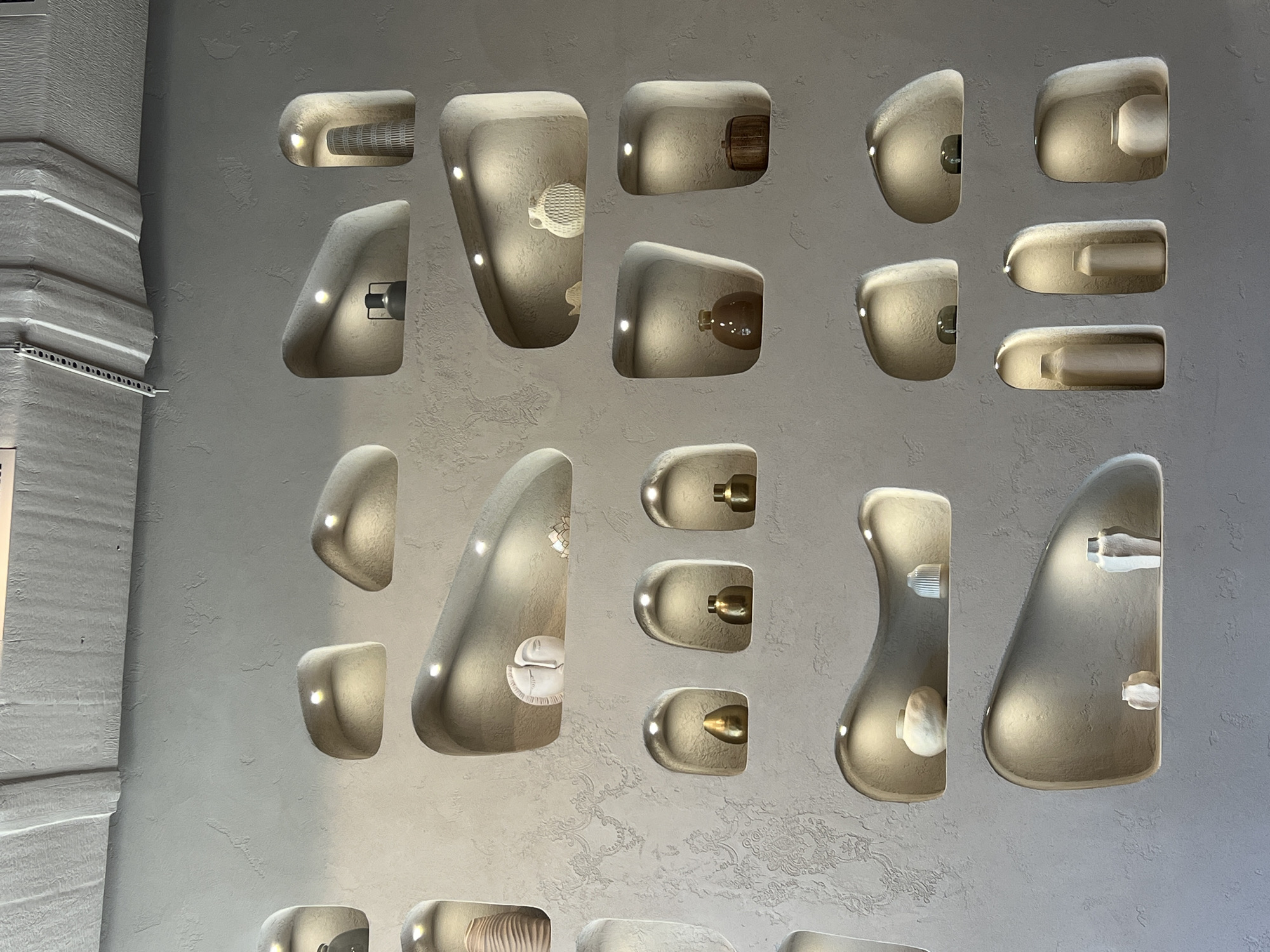 The image shows a close-up of a wall display at the TSUM (Central Department Store) in Odesa, Ukraine. The wall has several irregularly shaped recesses or niches, each illuminated from within. Various decorative items are placed inside these niches, including what appear to be vases, pots, and other small objects, some made of woven material. The wall itself has a textured, off-white surface. To the left of the display is a fragment of a wall with horizontal grooves or ridges.