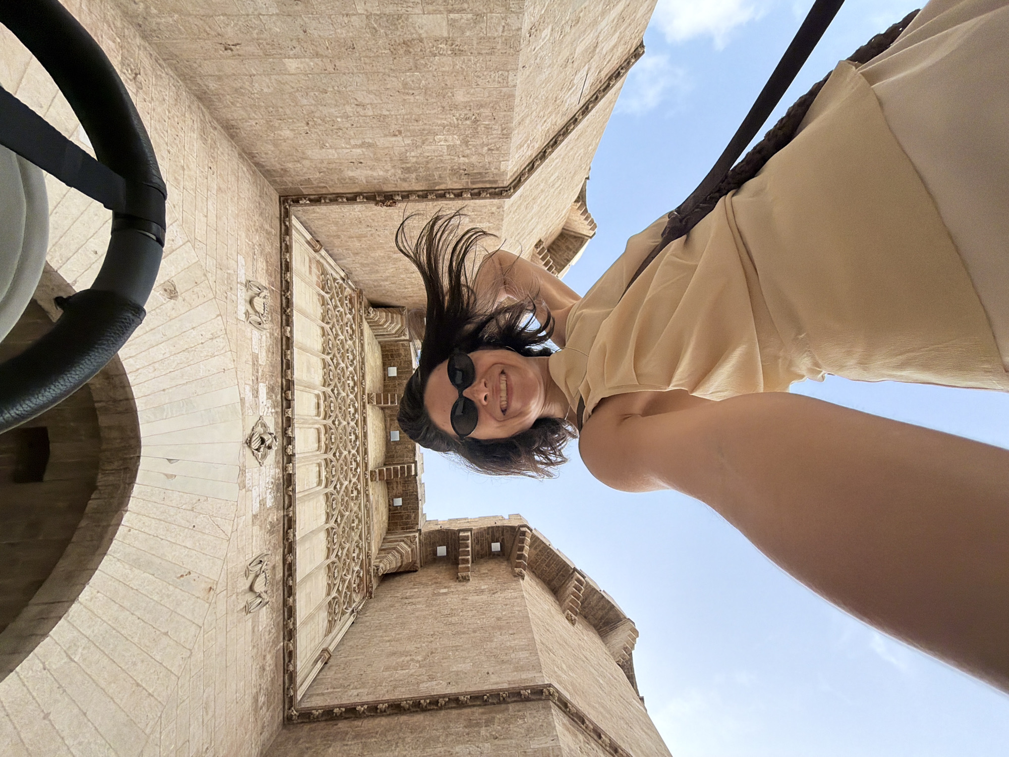 In a low-angle, wide shot, a woman with dark hair, sunglasses, and a beige dress smiles at the camera, holding the camera out in front of her. She is positioned below a stone archway, with the sky visible above. The architecture appears ancient, with ornate carvings and textured stone walls. A black strap is visible across her shoulder and extending out of frame. The scene is brightly lit by daylight.