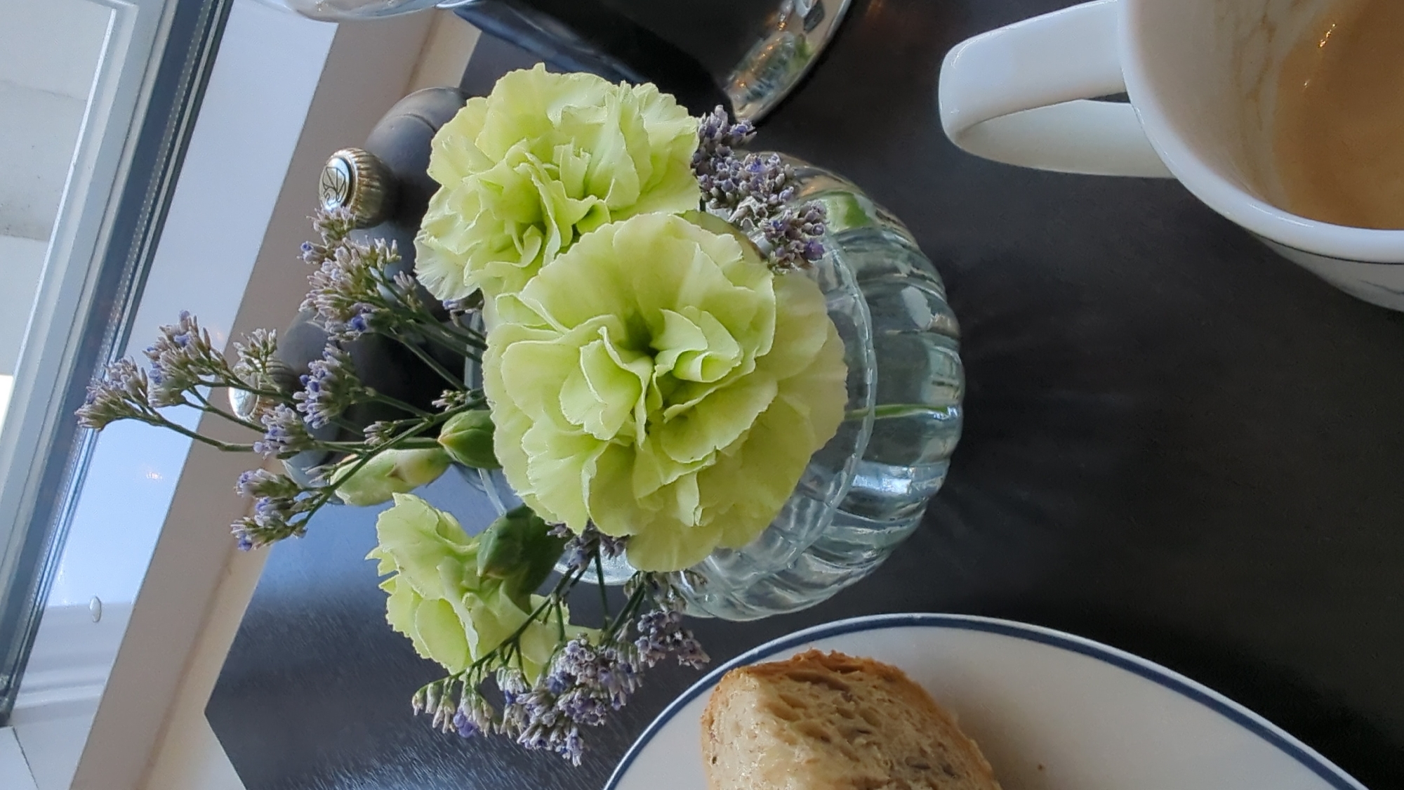 A vase of pale green carnations and purple statice is placed on a dark surface. To the left, a slice of bread is on a white plate with a blue rim. To the right, a white mug with a handle contains a beverage. The background is a window with a view of a brightly lit sky. The setting is a room in Larkollen, Norway.
