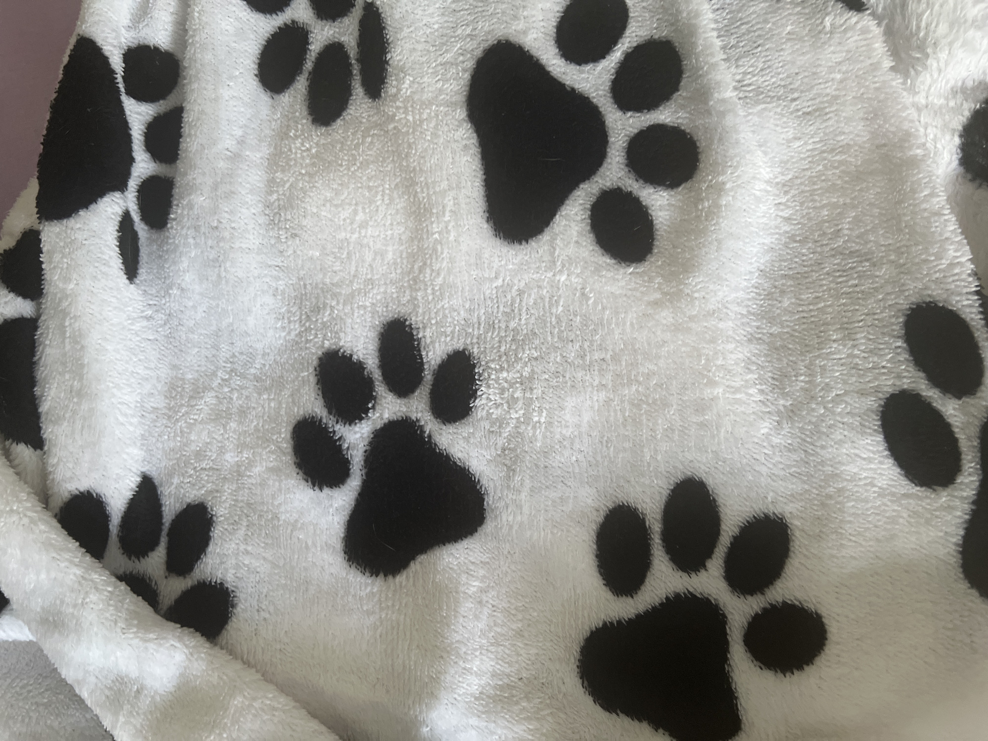 The image displays a close-up, top-down view of a white plush blanket with a pattern of black paw prints. The blanket is folded in places, creating shadows and texture. The paw prints vary in size and orientation, with some appearing complete and others partially obscured by the folds. The material of the blanket is soft and fuzzy, as indicated by the texture in the image. There is no identifiable location in the city of Krasnodar, Russia. There are no people, actions, or interactions depicted in the image.