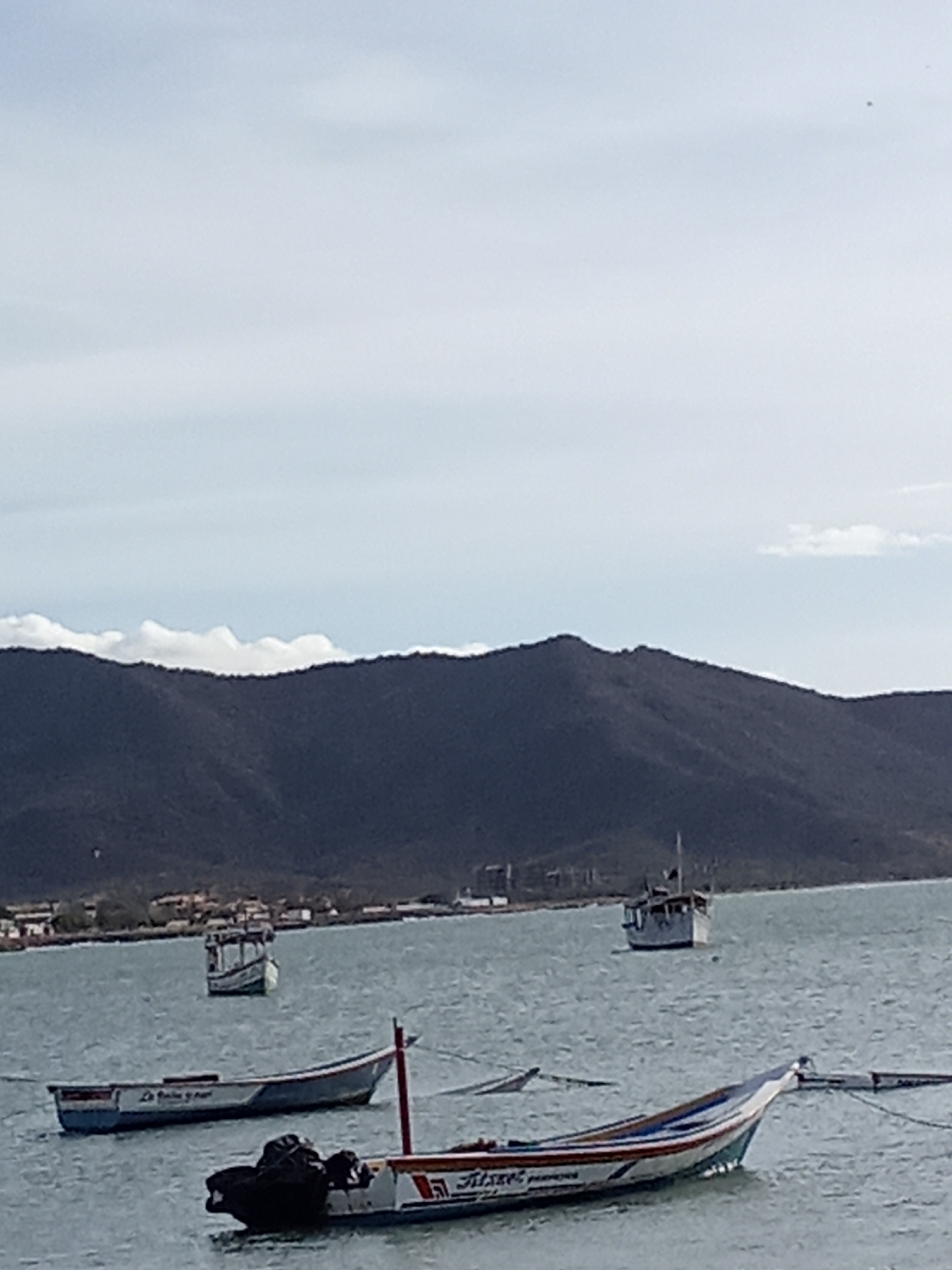 The scene depicts a coastal area in Juan Griego, Venezuela. In the foreground are three small boats moored in the water. One is closer to the viewer with what appears to be dark colored equipment covered up. A long slender fishing boat can be seen next to it and another further in the background, partially obscured by the other boats and closer to the land. In the midground, there is a mountain range along the coastline, with scattered buildings visible near the water's edge. The sky is overcast with some cloud cover.
