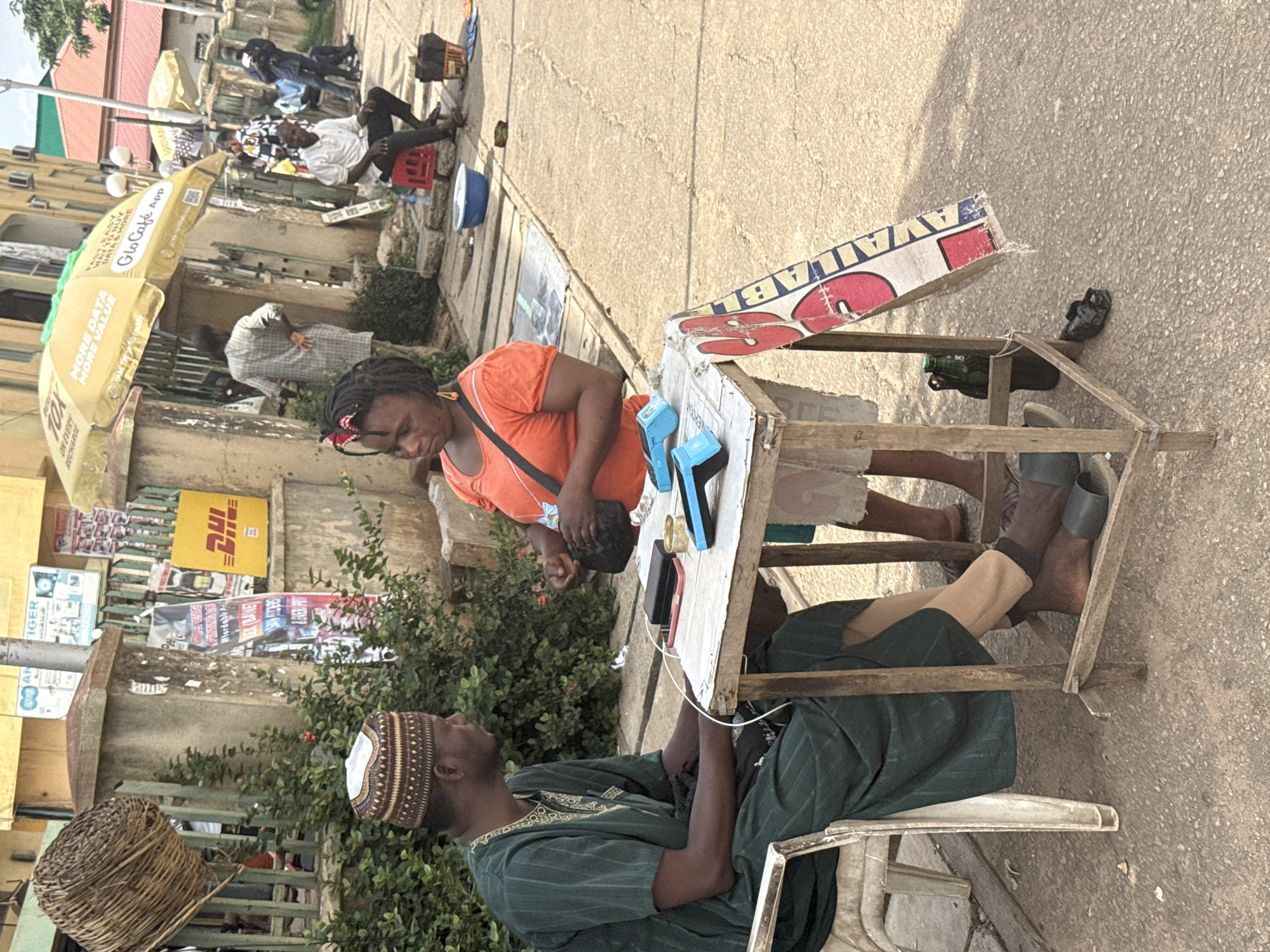 The image shows a woman in an orange shirt standing behind a makeshift wooden table, likely providing phone charging services to a man sitting in a chair in front of the table. The man is wearing a dark green robe and a traditional patterned cap. Cables and phones are visible on the table, indicating the charging setup. Overhead is a sign reading "AVAILABLE". The setting appears to be a street in Abuja, Nigeria, judging by the local advertising on the sides of the buildings, including banners for "Glo Cafe App" and "MORE DATA MORE VALUE". The context suggests a scene of informal economic activity in an urban environment.