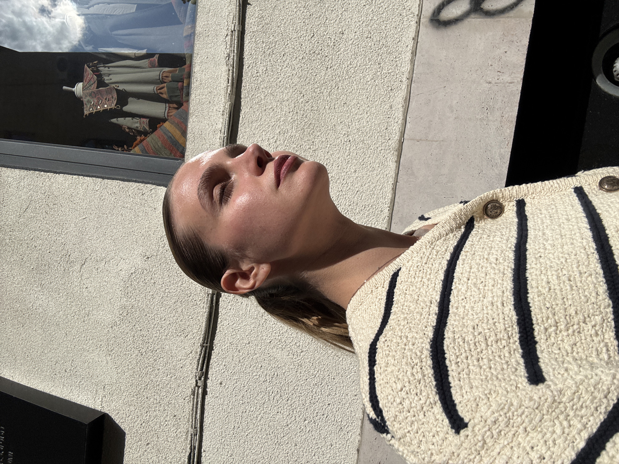 A medium close-up shot captures a woman in profile, positioned on her back and gazing upwards with her eyes closed, seemingly basking in bright sunlight. Her hair is pulled back, and her face shows a luminous sheen from the sun. She wears a cream-colored knitted cardigan with bold dark stripes and prominent gold-toned buttons.
The background reveals an urban setting. To the left, a textured, light-colored building wall features a window reflecting a partly cloudy sky. Inside the window, a display item, possibly a traditional patterned garment or umbrella in shades of green, red, and orange, is visible. Running vertically along the wall are thin electrical cables. Below the woman and extending towards the bottom left, a dark, angular object with faint, indistinct text (possibly "RECORD" or "PROD") sits in shadow on a concrete surface. Further right, faint, dark, illegible graffiti is visible on a lighter concrete area.
The strong, direct sunlight creates high contrasts and sharp shadows, indicating a clear, sunny day, likely mid-day. The woman's closed eyes and upward gaze convey a sense of peaceful relaxation. The scene is set in Tbilisi, Georgia.