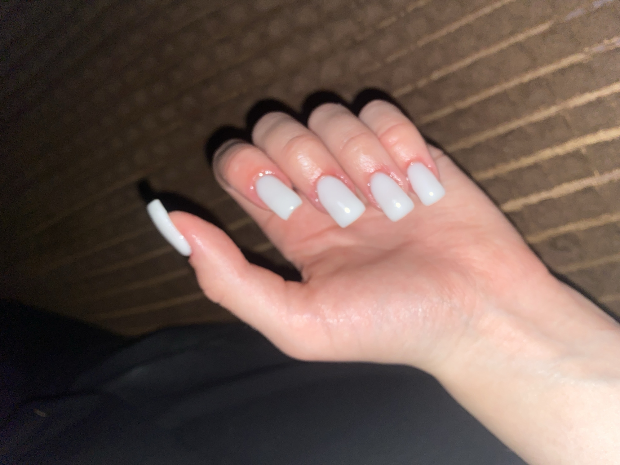 A hand with square-shaped, white acrylic nails rests on a brown, ribbed surface. The hand is positioned in the foreground of the image, with the fingers slightly curled. The background is dark and out of focus, with a hint of black fabric to the left and the ribbed surface continuing to the right. The scene is dimly lit, with a soft glow illuminating the nails. The location is not specified in the image.