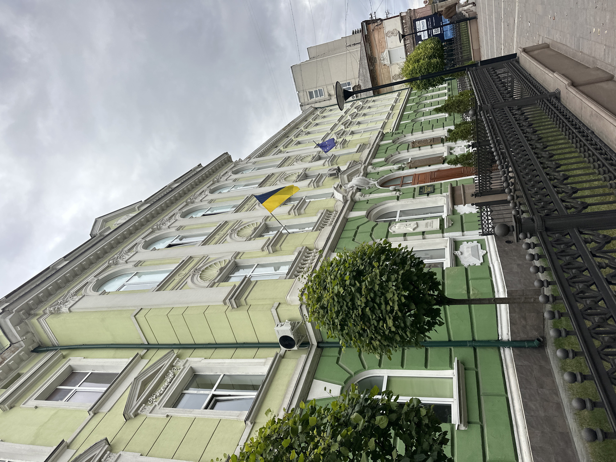 The image shows a street view of a historic building in Ternopil, Ukraine, identified as the Ternopil Regional State Administration. The building is painted a light green with white ornate trim around the windows and cornices. It has multiple stories with arched windows on the upper floors. A Ukrainian flag and a European Union flag are visible flying from the building. In front of the building, there is a black wrought-iron fence. Two trimmed trees are planted in front of the fence. The street pavement is visible, and a lamppost stands to the right of the building. In the background, other buildings and a cloudy sky are present. A person is seen walking on the sidewalk in the distance.