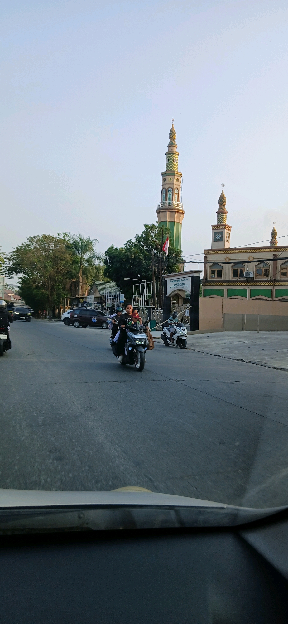 In Samarinda, Indonesia, a street scene unfolds with a prominent mosque on the right, featuring a tall green and gold minaret and an adjacent building with similar architectural elements. The sky is a clear, pale blue, suggesting it's either early morning or late afternoon. In the foreground, a gray asphalt road is visible, with visible cracks and markings. Traffic includes several motorcycles, with one carrying three people in the center of the frame: a driver and two passengers. Another motorcycle is visible to the right, and one to the left. Cars are also present on the road, including a dark-colored vehicle and a lighter-colored one. Trees line the left side of the road, with a prominent palm tree among them. The overall impression is a typical urban street environment in Indonesia during daylight hours.