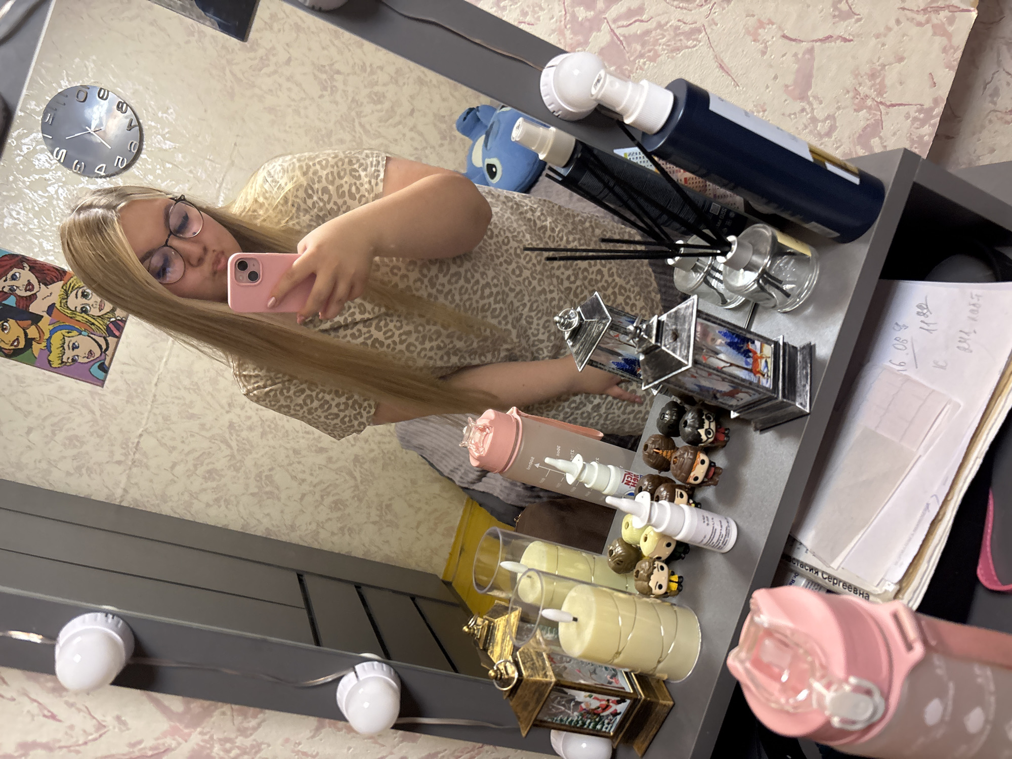 The image depicts a young woman with blonde hair, wearing glasses and a leopard-print top, taking a selfie in a mirror. She is holding a pink smartphone. The mirror reflects a shelf filled with various items, including decorative lanterns, candles, bottles of liquid, and small figurine characters. In the background, a clock and framed artwork are visible on the wall. The overall setting appears to be a room, possibly a vanity or dressing area. There is no indication of the location being in the city of Большой Букор, Russia.