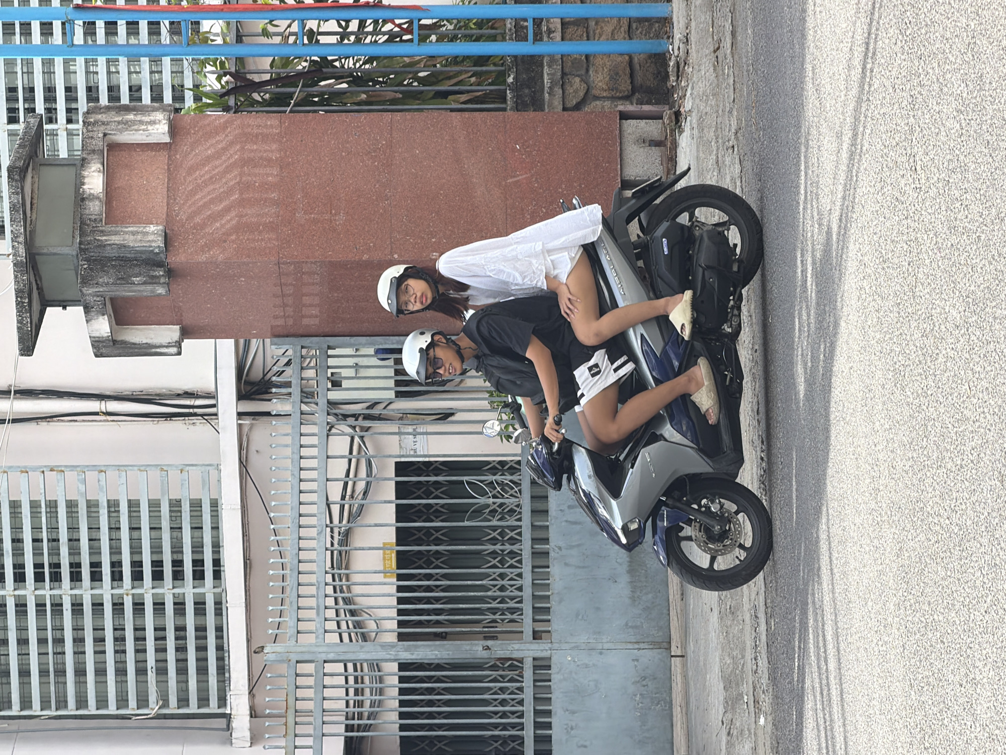 Two young people, a male driver and a female passenger, wearing helmets ride a blue and silver scooter on a paved street in Nha Trang, Vietnam. The male driver wears a black t-shirt and white shorts with black trim. The female passenger wears a white, long-sleeved, voluminous dress and white slide sandals. Both are wearing white helmets. The scooter is moving from left to right. In the background, there is a large, ornate gate made of grey metal bars. Behind the gate is a building with metal louvers covering windows and a red granite pillar with a concrete structure on top. The scene appears to be a street in a residential or commercial area of Nha Trang.