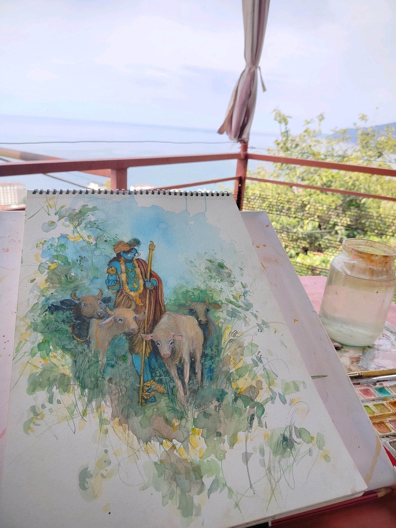 The image shows a watercolor painting of Lord Krishna with cows, resting on a table outdoors. In the background, there is a railing, trees, and the sea. The painting is on a spiral-bound sketchbook, and a jar of water and a watercolor paint palette are also visible on the table. The overall scene suggests an artist at work in a scenic location. The location appears to be Gagra, Abkhazia, given the distinct architecture of the railing and the style of the building visible in the distance.
