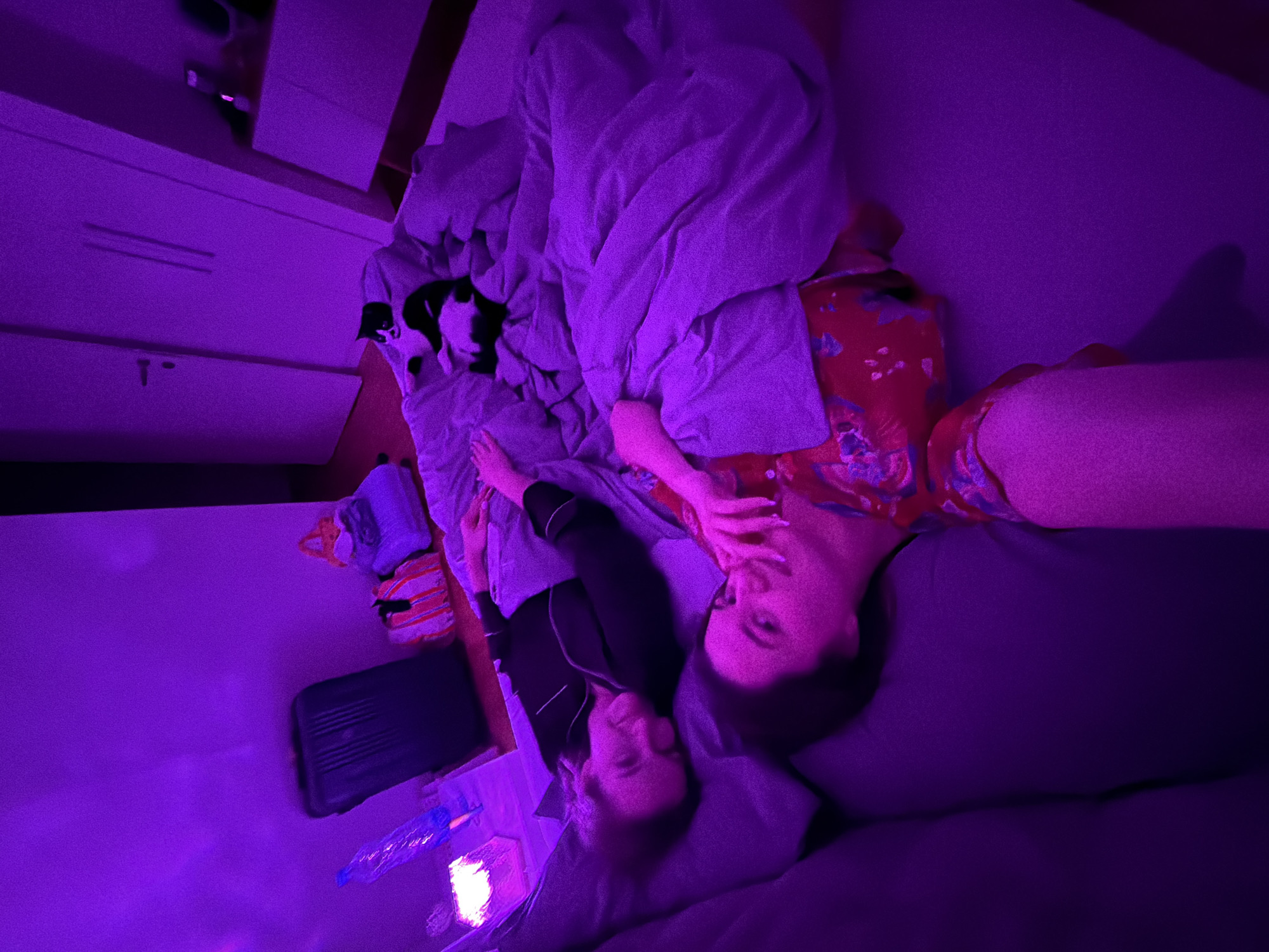 A photograph captures a woman in a floral patterned garment lying on a bed, next to another woman wearing a black shirt. A cat is partially visible under the duvet. A white dresser is located in the background. In the foreground, a suitcase and plastic water bottle are visible. The image is illuminated in purple hues. It is not possible to determine if the location is in Belgrade, Serbia.