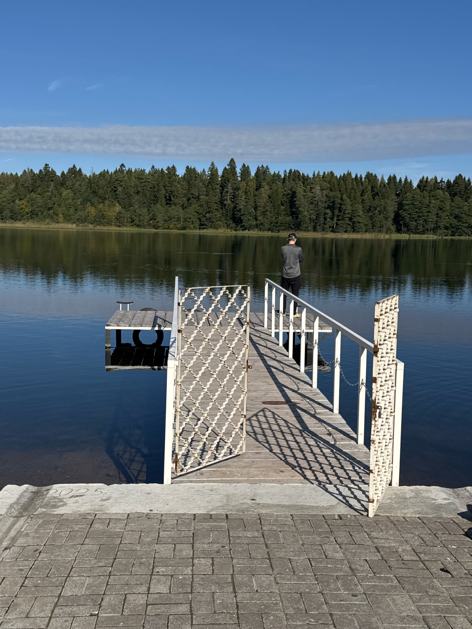 The image depicts a person standing on a wooden pier that extends into a calm body of water. The pier has white railings and a decorative white gate that is open. A smaller, separate wooden platform with a tire and a seat is visible to the left of the main pier. The water reflects the clear blue sky, which has a thin band of clouds. Across the water, a dense forest of evergreen trees lines the shore. The foreground shows a paved area made of grey interlocking bricks leading up to the concrete edge of the pier. The number "2025" is faintly visible etched into the concrete. The scene appears to be a serene outdoor setting, possibly a lake or large pond, in a natural environment. The location is identified as Unknown, Russia.