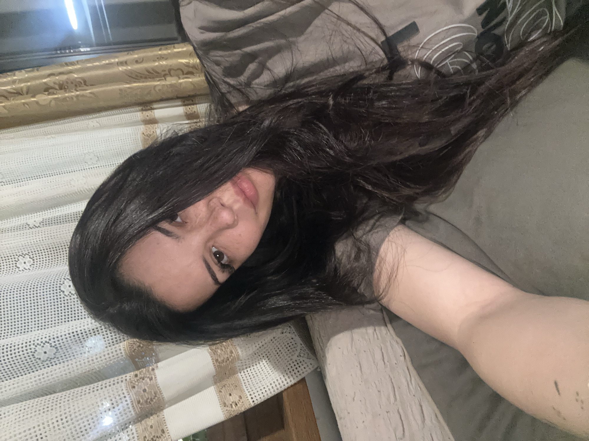An indoor, medium close-up image shows a person, likely female, with long, dark hair, lying down on what appears to be a bed or sofa, looking directly at the camera. The image is oriented diagonally, with the person's head towards the lower left and their body extending towards the upper right.
The subject's face is partially visible, featuring dark eyes, dark eyebrows, and slightly parted lips with a natural or light pink hue. Their long, dark hair cascades around their head and shoulders, blending with the dark grey or brown bedding. A bare arm is visible in the lower right, resting on the same surface.
In the background, to the left of the subject, there is a window covered with two layers of curtains. The inner layer is a white, delicate lace curtain with floral patterns. Behind it, a thicker, patterned curtain in a golden-beige color with ornate designs is visible. A dark, reflective surface, possibly a window pane reflecting an indoor light source, is in the top left corner. A dark wooden frame or furniture edge is also visible beneath the curtains.
On the dark grey pillow or bedding near the person's head, in the upper right quadrant of the image, there are white or light grey stylized graphic elements that resemble abstract lettering or a logo, with discernible shapes that could be "WE" or similar characters, alongside wavy lines.
The overall impression is one of a relaxed, informal moment, likely a selfie or a casual photo taken in a home setting, possibly during the evening due to the indoor lighting and lack of external light cues. The location context is Chubra, Bulgaria.