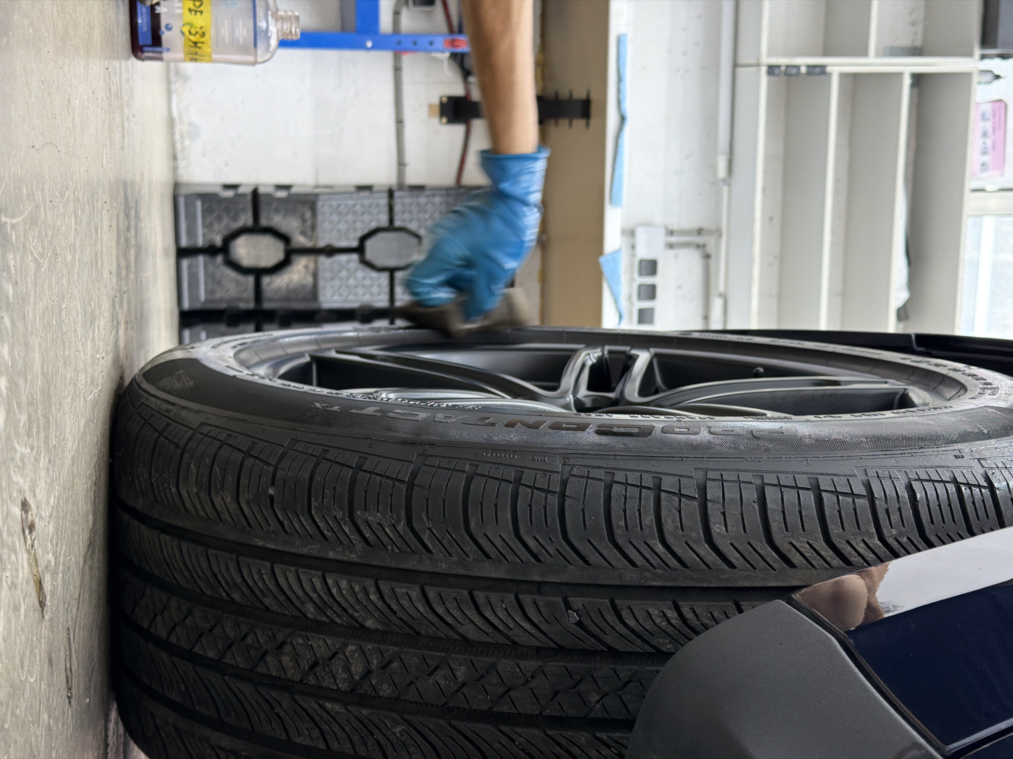 A low-angle, close-up shot shows a person wearing a blue glove cleaning the side of a car tire with a cloth. The tire is black and has a tread pattern. The car is dark blue. In the background, there are white shelves, a blue rack, and a bottle of liquid with yellow lettering that appears to say "РЕЗИНА" (rubber). The scene is indoors, on a concrete floor, suggesting a garage or workshop environment. The specific location in Krasnoyarsk, Russia, cannot be determined from the visual information provided in the image.