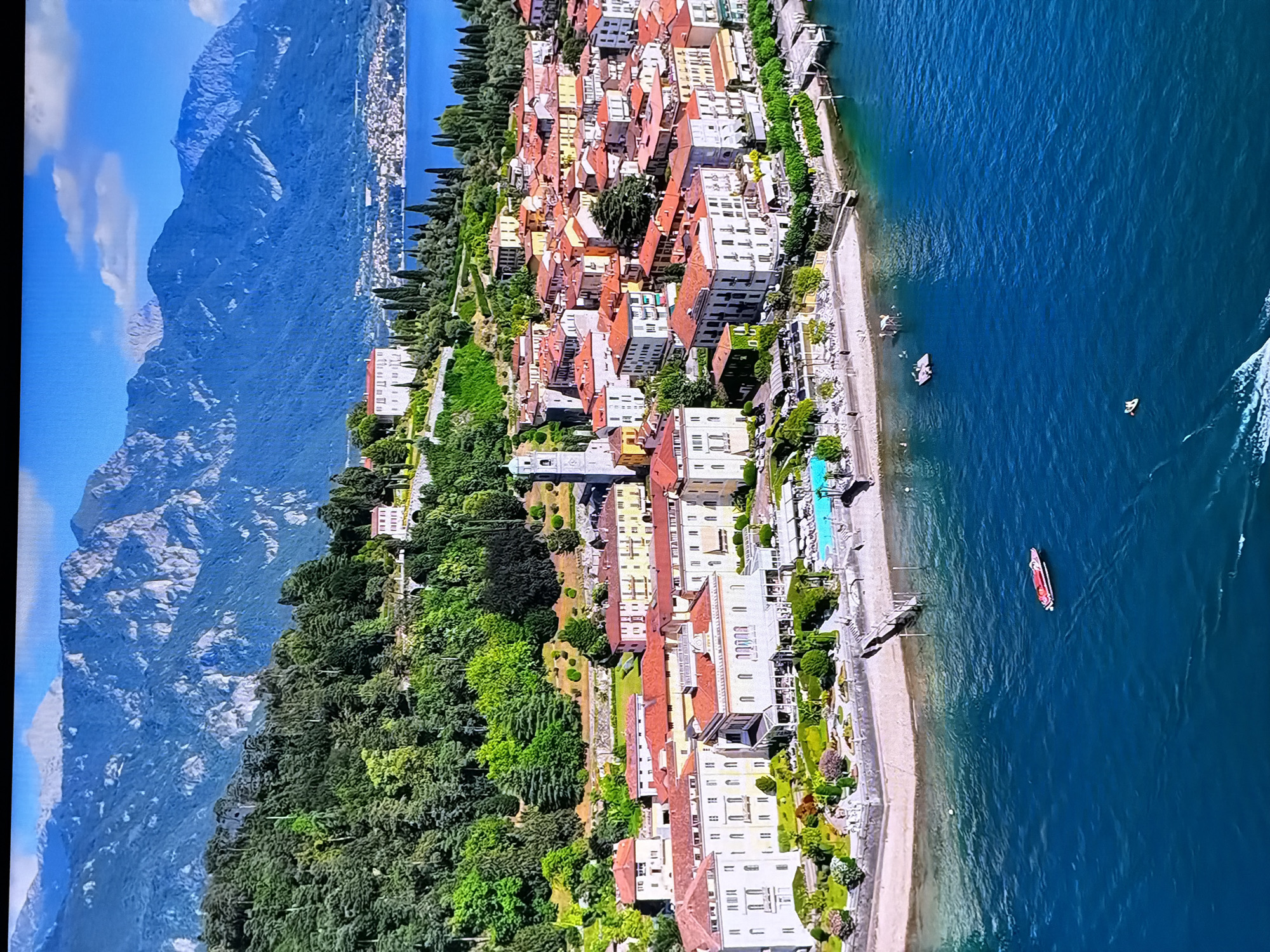This is an aerial view of a picturesque lakeside town nestled against a backdrop of lush green mountains. The town features a dense cluster of buildings with terracotta roofs, suggesting a European architectural style. A prominent church tower with a clock is visible amidst the buildings. In the foreground, the deep blue water of a lake or sea is visible, with a small red and white boat in the center. Along the shoreline, there is a sandy beach area with lounge chairs and a swimming pool, indicating a resort or hotel. The surrounding hills are covered with dense green trees, and the distant mountains are rugged and rocky, with some snow-capped peaks visible. The sky is a clear blue with scattered white clouds. The scene depicts a serene and beautiful landscape, ideal for tourism.
The location is NOT in the city of Chirchik, Uzbekistan. This image depicts a European lakeside town, likely in Italy given the architecture and the style of the mountains and lake.
