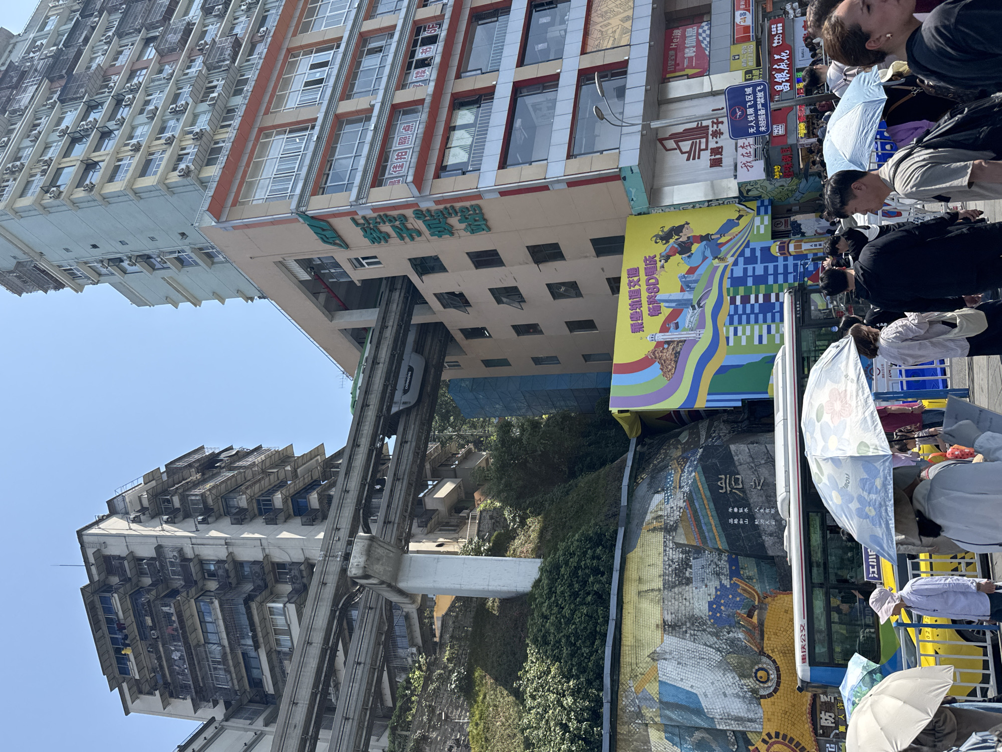 The image shows a scene in Yuzhong District, China, featuring the Liziba Station. The elevated train track runs through the middle of buildings. There are buildings on both sides, with lots of air conditioning units. Crowds of people are seen on the right side of the image, some holding umbrellas. Signage and advertising are visible, including promotional material for Chongqing transportation and local businesses. A mosaic wall is also present. The sky is clear and blue.