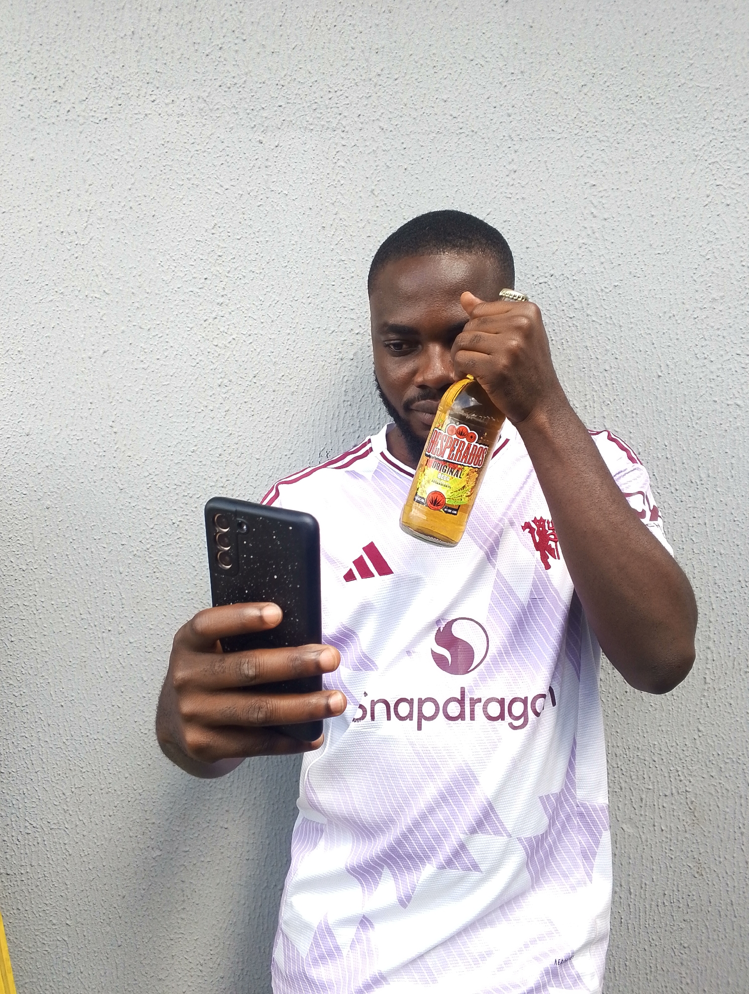 A young Black man stands in front of a textured gray wall, holding a smartphone in his left hand and a bottle of Desperados Original Beer in his right. He is wearing a white and purple soccer jersey with the Adidas logo and the Manchester United crest visible. The Snapdragon logo is also prominent on the jersey. The man is looking at his phone, suggesting he might be taking a selfie or recording a video. The lighting indicates it is daytime, possibly outdoors due to the bright natural light. The man has short hair and a beard, and is wearing rings on his fingers. The bottle of Desperados beer is yellow and appears to be partially full. The overall mood of the image is casual and relaxed. The setting is likely urban, given the wall and the style of clothing.