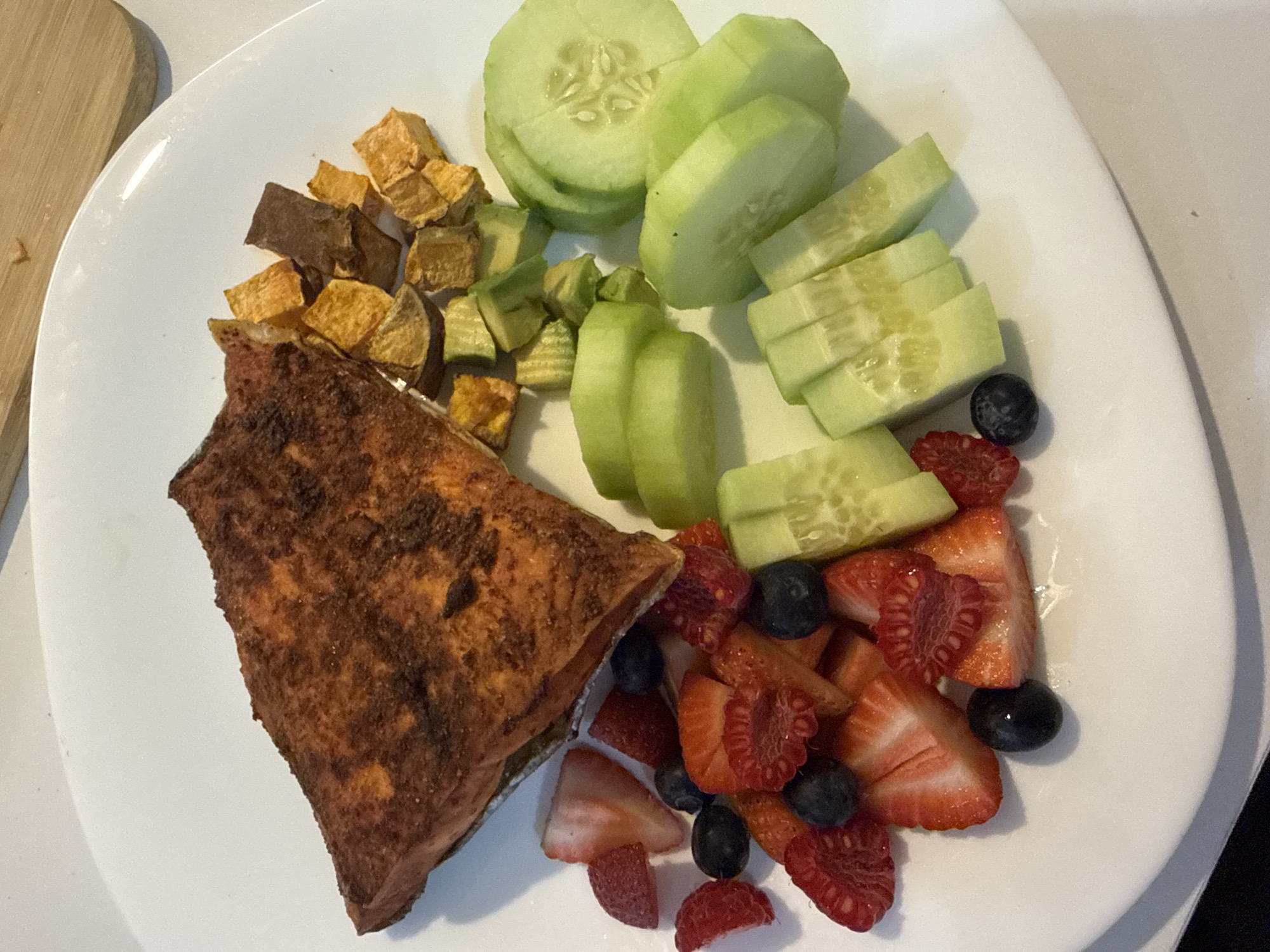 A brightly lit, overhead shot showcases a healthy meal served on a white, oval plate. The central focus is a substantial piece of seasoned salmon, appearing baked or pan-fried, with a browned and textured surface, positioned towards the lower left.
Adjacent to the salmon, in the upper left quadrant of the plate, are cubed and roasted vegetables, likely sweet potatoes or possibly butternut squash, displaying varying shades of orange and brown. To their right, a scattering of diced green avocado pieces is visible.
A generous portion of fresh cucumber occupies the top and middle sections of the plate, presented in various cuts including thick rounds, half-moons, and smaller rectangular slices.
The lower right section of the plate is filled with a colorful mix of fresh fruit, comprising sliced red strawberries, whole or halved red raspberries, and dark blue blueberries.
A wooden cutting board is partially visible in the top left corner, suggesting an indoor setting like a kitchen or dining area. The image captures the meal as if ready to be eaten. There are no people visible, nor is there any discernible text. While the location context is provided as Puebla, Mexico, there are no visual cues within the image itself to confirm this geographical detail.