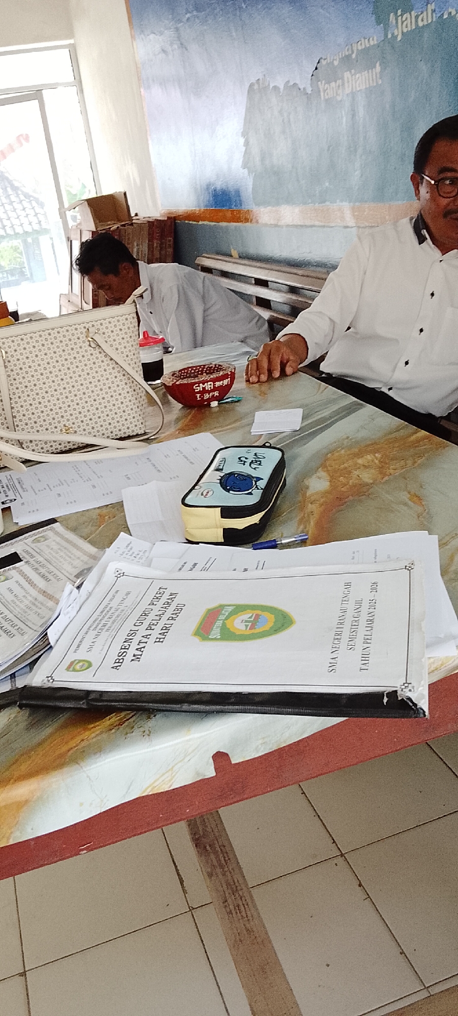 The image shows two men sitting at a table, likely in a school or office setting. One man, wearing glasses and a white collared shirt, is seated on the right, looking towards the camera. The other man, wearing a white shirt, is seated on the left, looking down at the table.
On the table, there are several documents, including a binder labeled "ABSENSI GURU PIKET MATA PELAJARAN HARI RABU" which translates to "Teacher's Duty Roster Subject Wednesday". There's also a pencil case with a cartoon character and a red bowl with writing that appears to be "SMA NEGERI I-BPR". A white patterned bag is also visible on the table. The background features a wall with a blue and white mural and a wooden bench. The overall environment suggests an indoor workspace. The image does not contain enough information to determine its specific location within Jepara, Indonesia.