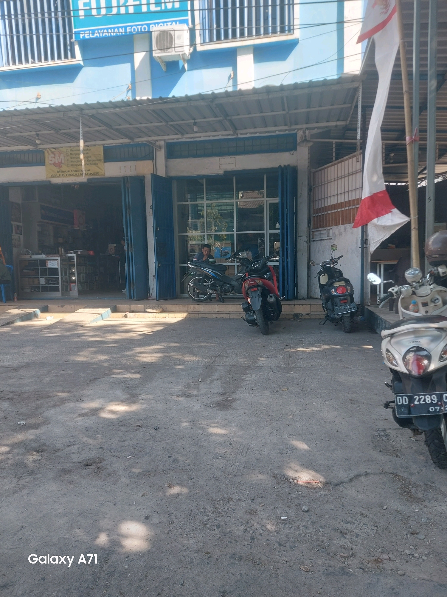 A low-angle, eye-level medium shot shows the exterior of a Fujifilm photo service shop in Makassar, Indonesia. The shop has a blue facade with "FUJIFILM PELAYANAN FOTO DIGITAL" signage. A metal awning covers the entrance, which is open, revealing shelves of products inside. Three motorcycles are parked in front of the shop: a red scooter, a black scooter, and a white scooter. The red and black scooters are parked in the driveway, while the white scooter is parked on the sidewalk to the right. A man is sitting on the black scooter, looking to the left. A red and white Indonesian flag is displayed on a pole to the right of the shop. The ground in front of the shop is made of asphalt and dirt, with shadows from trees dappled across it. The overall scene is bright and sunny.