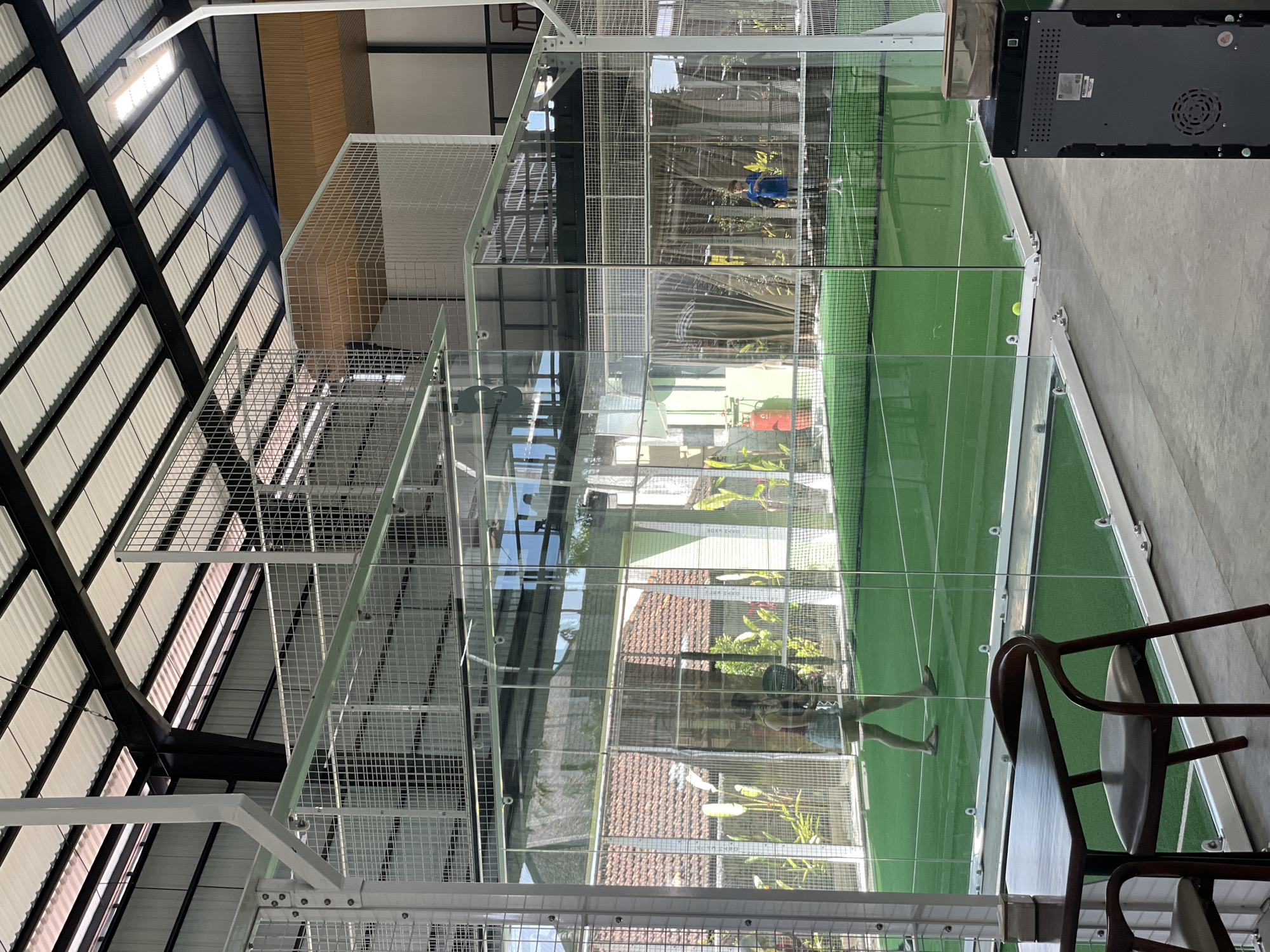 In Kuta Selatan, Indonesia, a padel tennis court is situated indoors with a high, corrugated metal roof supported by black metal beams and white structural elements. The court is enclosed by clear glass walls and a green mesh net, with a bright green turf playing surface marked by white lines. Two individuals are visible on the court: a woman in a red tank top and denim shorts on the left, holding a padel racket, and a young boy in a blue t-shirt and grey shorts on the right, also with a racket. Outside the court, in the foreground, a dark wooden table with chairs is set up. Adjacent to the court, a black electronic device with vents and a small screen sits on the ground. The background outside the glass reveals a glimpse of tiled roofs and lush green vegetation, indicating a tropical environment.