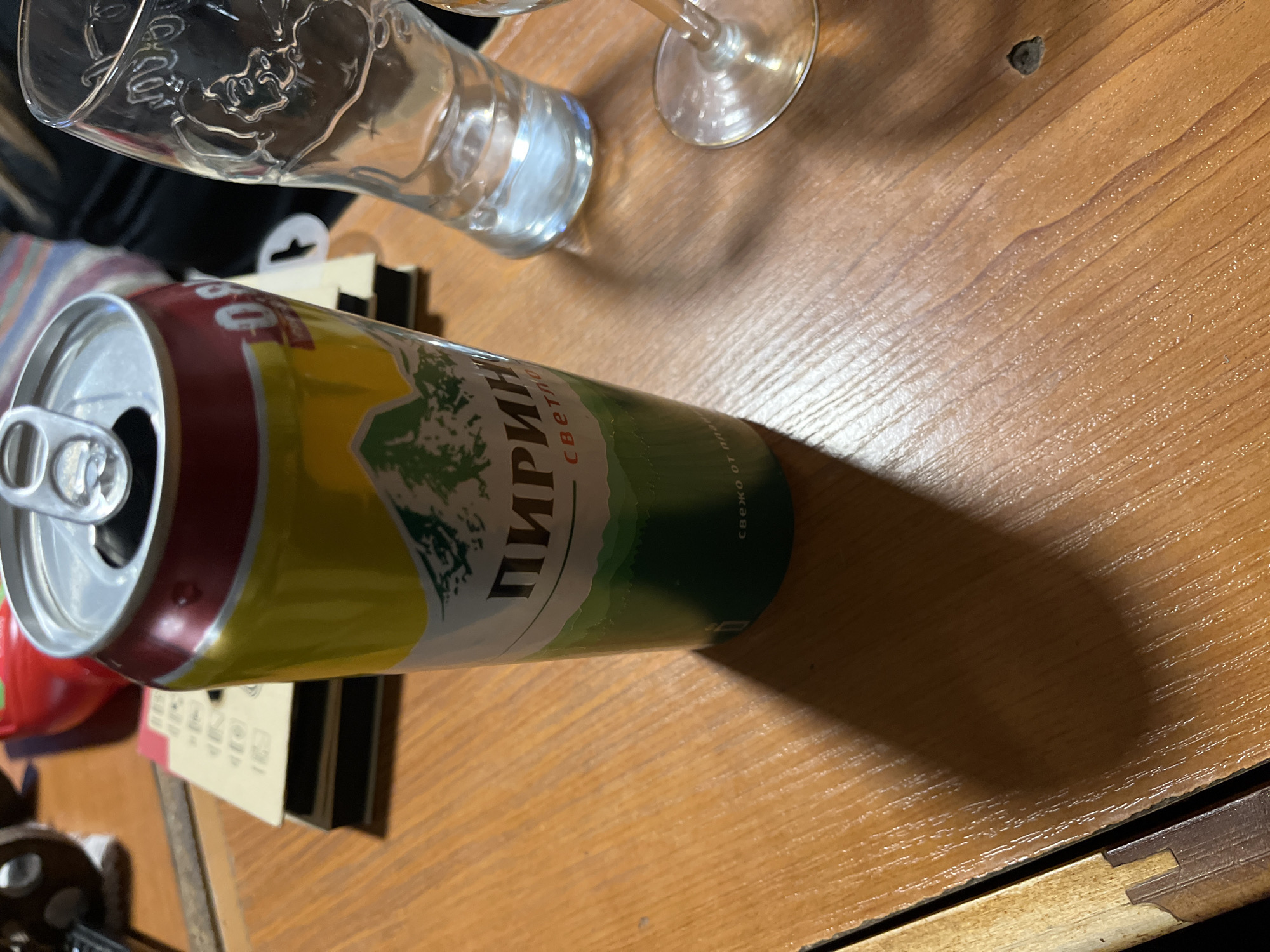 The image depicts a can of "Pirin Svetlo" beer lying horizontally on a wooden surface. The can has a red top, a yellow middle section, and a green lower portion. The can is open. Two empty glasses are near the can. The picture was taken indoors. The city Shopite, Bulgaria is not present in this image.