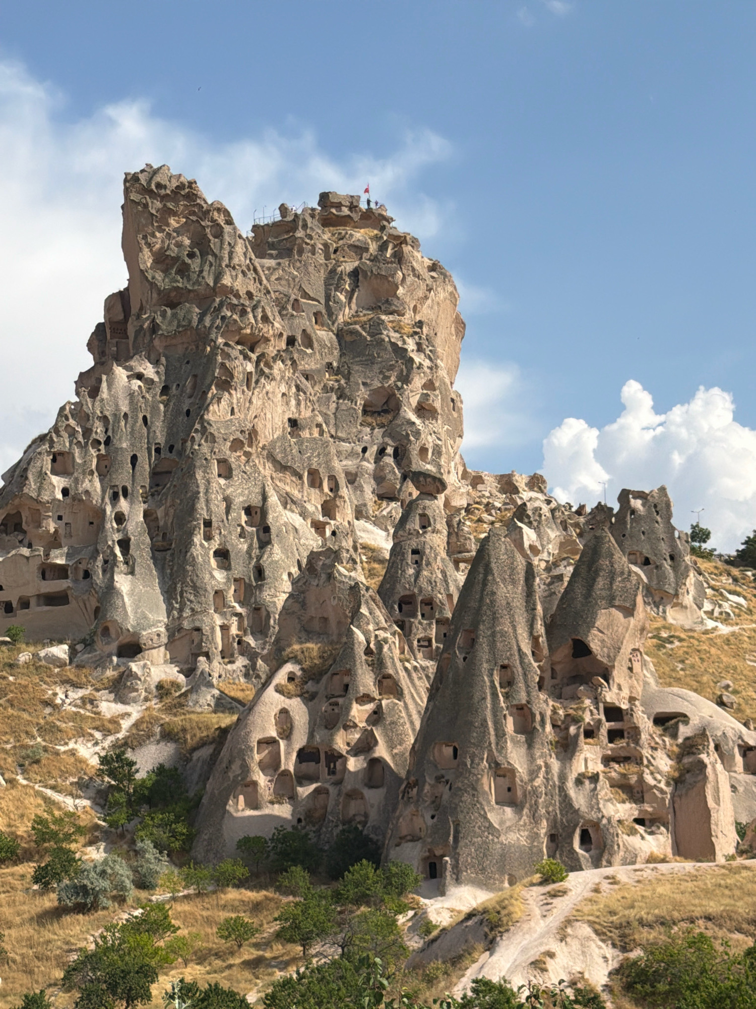 The image shows Uchisar Castle in Uchisar Beldesi, Turkey, a natural fortress carved into a large rock formation. The castle is a complex structure with numerous caves and openings, likely once used as dwellings, storage, and defensive positions. The rock formation rises prominently from a dry, hilly landscape dotted with sparse green trees and shrubs. The sky above is blue with scattered white clouds. Several people are visible near the top of the castle, appearing to be tourists exploring the site. The overall scene is a testament to the unique geological formations and historical human adaptation of the Cappadocia region.