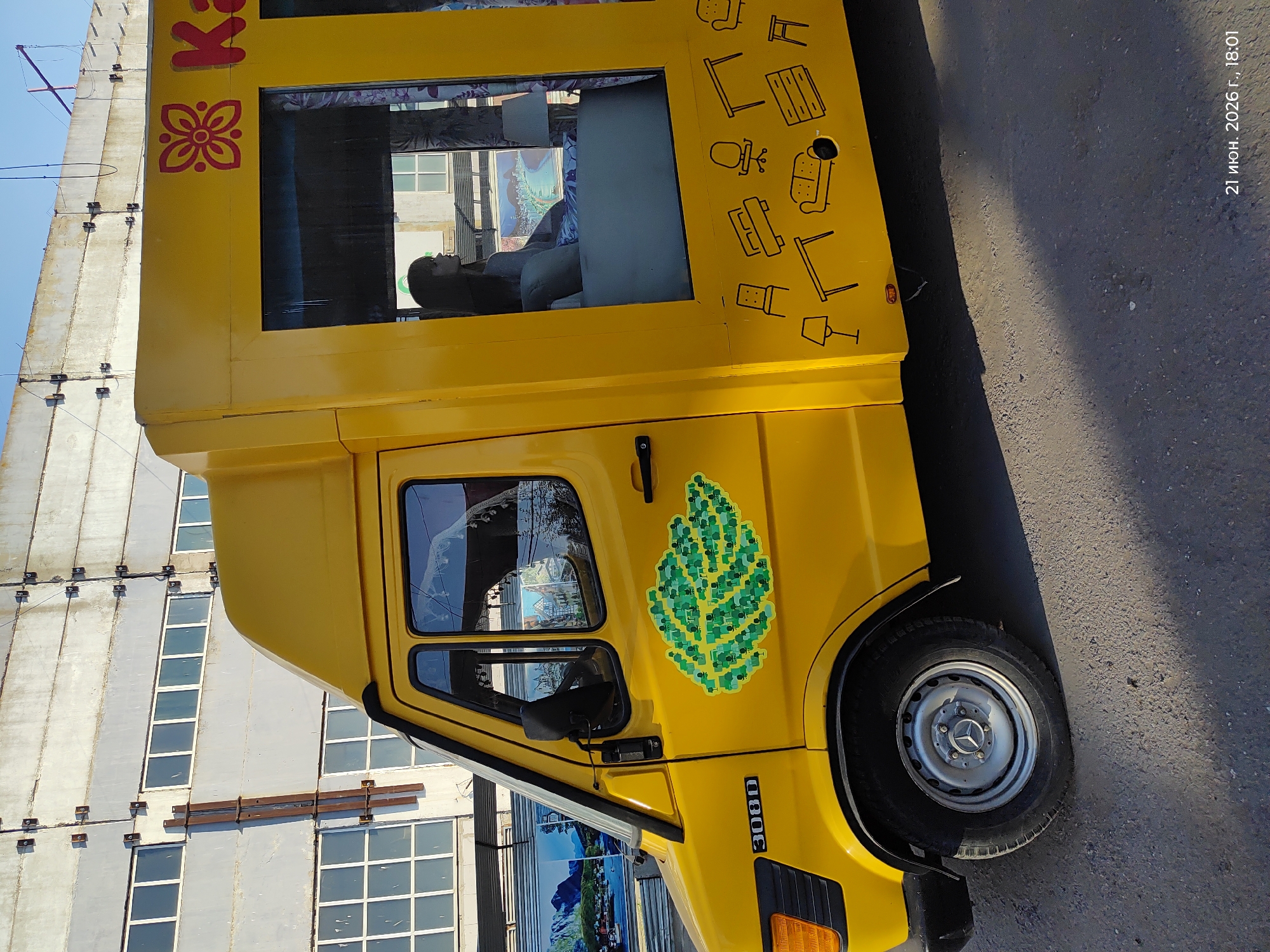 The image shows a bright yellow truck parked on a paved surface. The truck has several windows, one of which shows a person inside. The side of the truck is decorated with a stylized green leaf emblem and a black outline of furniture such as a desk, chair, and sofa. The truck's model number "308 D" is visible on the side. In the background, a large, grey, industrial-looking building with many windows is visible. The sky is blue, suggesting daylight. The overall impression is that of a commercial vehicle, possibly used for transporting goods or as a mobile service. A timestamp "21 июня. 2026 г., 18:01" is visible in the upper right corner, indicating the date and time the photo was taken. The lighting suggests it is late afternoon or early evening. The location is identified as Astana, Kazakhstan.