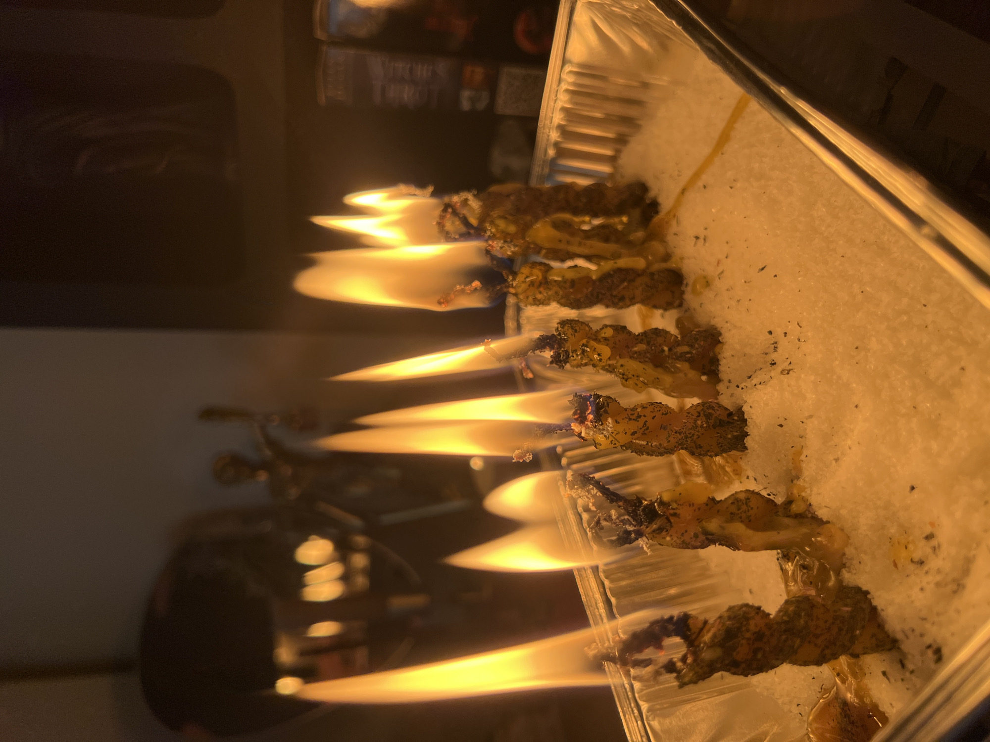 In the image, a line of small, twisted objects are burning, set in a clear container filled with a white granular substance. The objects appear to be some sort of incense or herbal material, with visible dried herbs and spices. They are actively burning with bright yellow and orange flames that cast a warm glow.
The background is dark and out of focus, but hints of objects can be seen. To the left, a dark, reflective surface suggests a piece of furniture or equipment. Further back, a blurred image of what looks like a bookshelf or display shelf contains some items, one of which has text that appears to say "WITCHES TAROT". This suggests a setting that might be a home or a shop related to spiritual or mystical practices.
The lighting is dominated by the flames, creating a dramatic and somewhat mysterious atmosphere. There are no people visible in the image. The overall impression is one of a ritualistic or decorative burning of incense. The granular substance appears to be salt or sand, often used in such practices. The image is taken from a slightly elevated angle, looking down at the burning items. There are no clear indications of the time of day or weather, but the indoor setting is implied.
