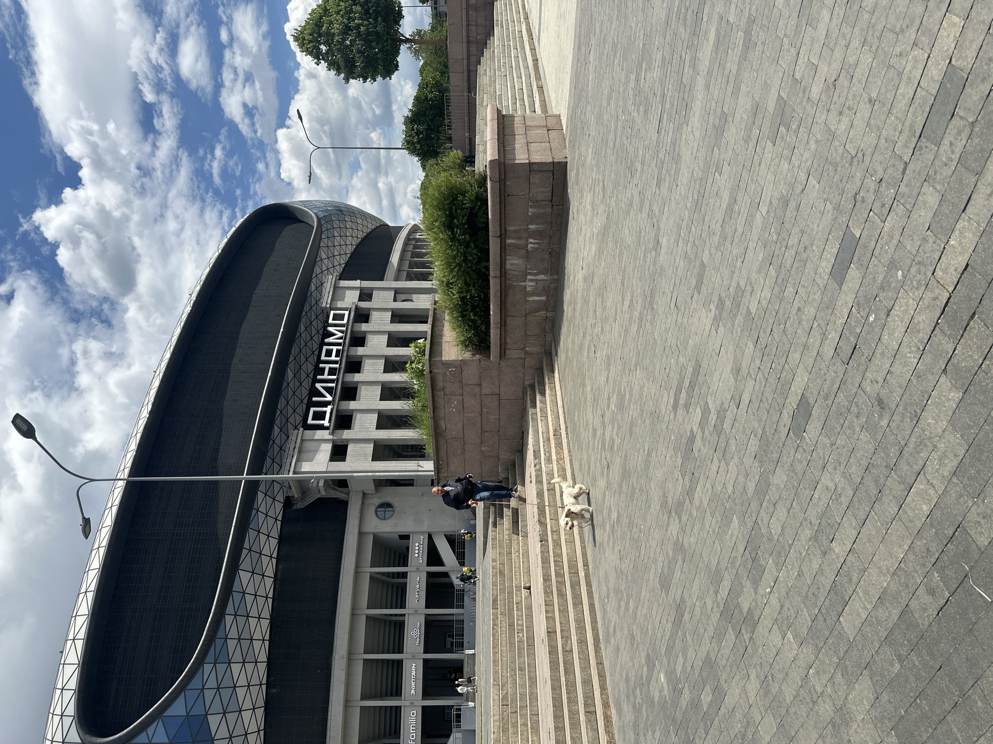 A man walks his small white dog down a wide outdoor staircase in front of a large modern stadium. The stadium has a curved, dark facade with geometric patterns and features a prominent "DYNAMO" sign in large white letters. The man is wearing a dark jacket and jeans and is holding a red leash. The dog is walking a few steps ahead of him. The staircase is made of stone blocks and is flanked by stone retaining walls with greenery on top. The ground in front of the stairs is paved with grey interlocking stones. In the background, there are trees and a bright blue sky with scattered white clouds. Several streetlights are visible, two near the stadium and one further in the distance. The overall scene is one of a bright, sunny day in an urban public space.