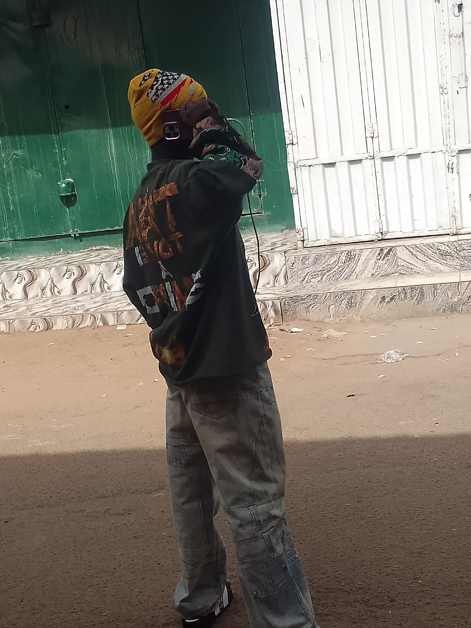 A person stands in Gombe, Nigeria, holding a phone to their ear. They wear a yellow and white patterned beanie, a black graphic sweatshirt, and light blue jeans. The ground appears to be a street. Behind the person are a green corrugated metal wall and a white metal door. The person appears to be having a conversation on the phone.