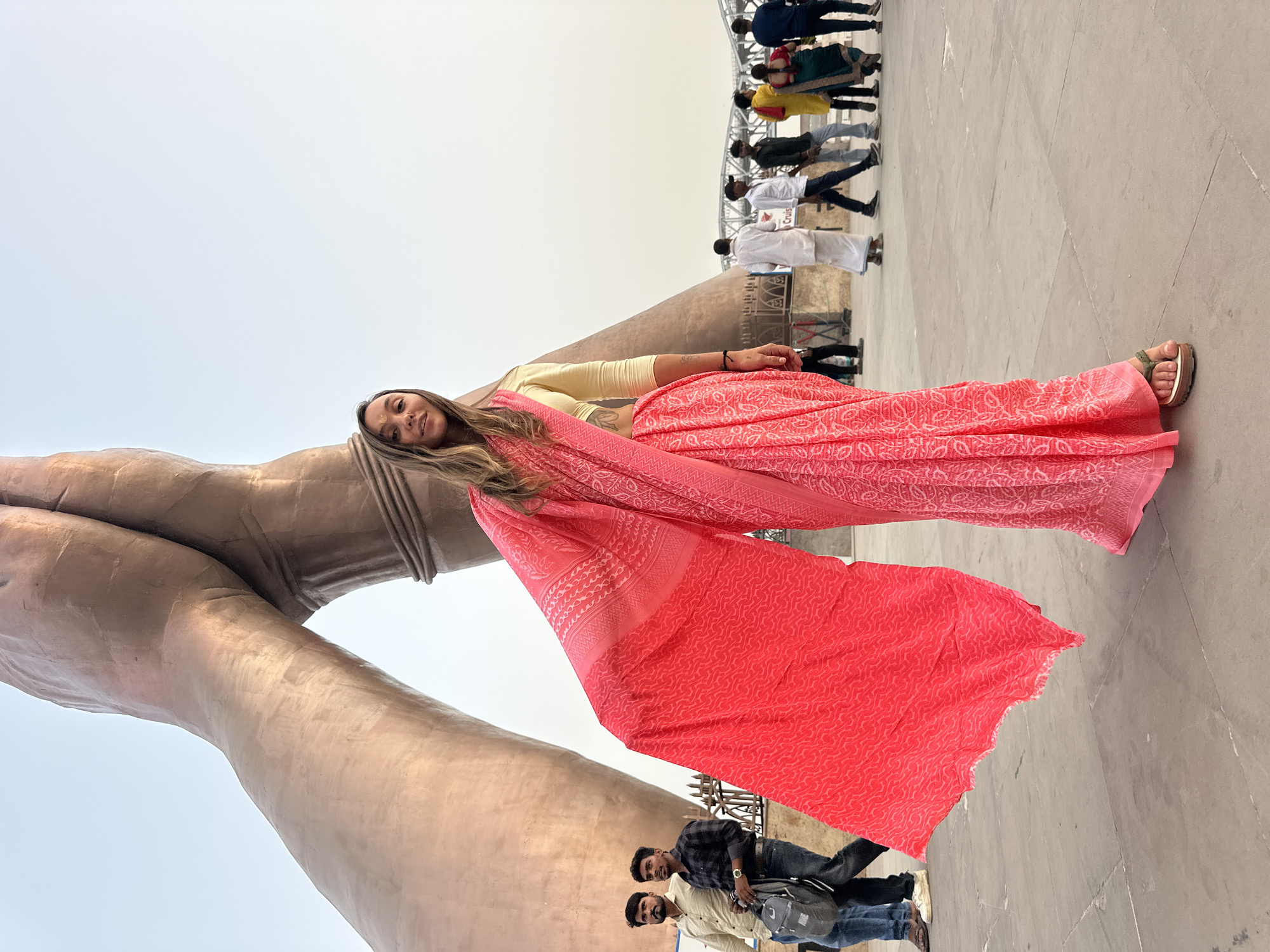 A woman is posing at a statue in Mughalsarai, India. The statue is a large pair of folded hands made of bronze. The woman is wearing a pink sari and a light yellow blouse. She is standing to the right of the statue and the sari is draped over the arms. In the background are other people standing and walking around on the platform.