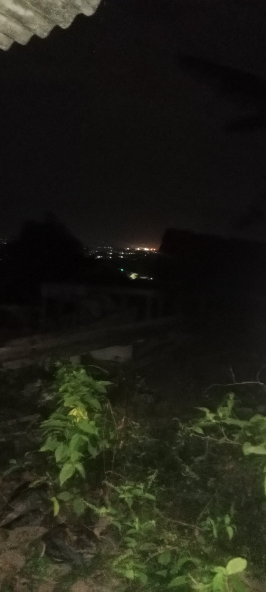A dimly lit night photograph captures a distant view of a city, likely Soko, Indonesia, characterized by a scattered pattern of illuminated buildings. The foreground is dominated by dark foliage and the silhouette of a corrugated roof, suggesting an elevated viewpoint. The sky is predominantly black, with only faint hints of celestial light. The scene appears to be viewed from a rustic vantage point, possibly a hillside or a rooftop, overlooking a sprawling urban landscape at night.