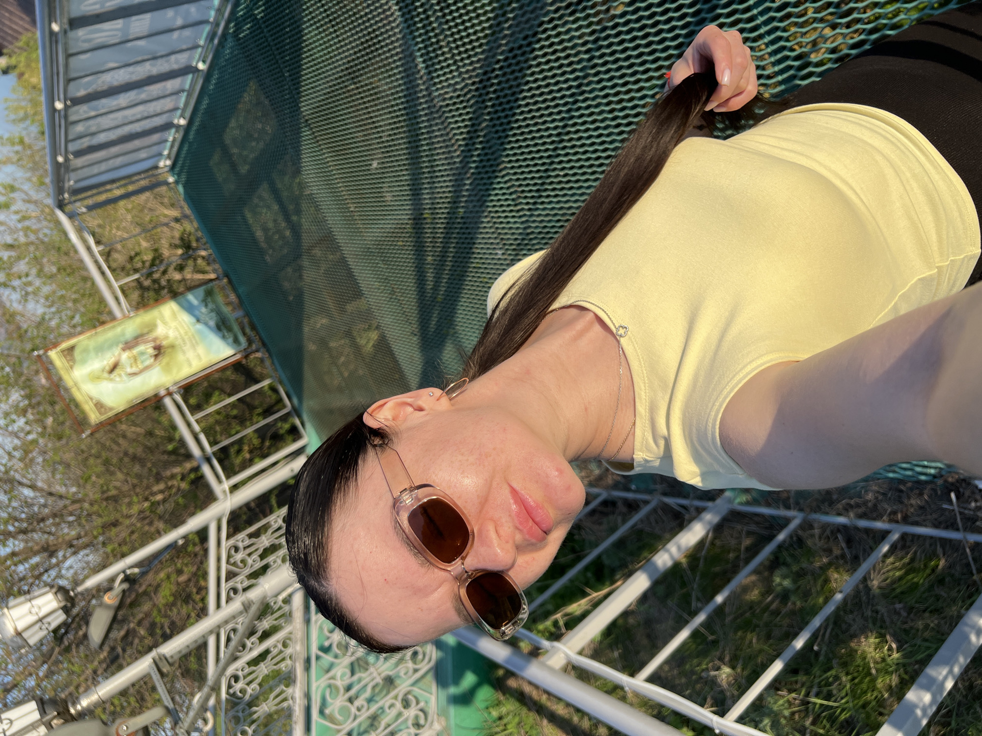 A person is positioned on a metal latticework platform or structure, likely an observation point, in what can be identified as Sviyazhsk, Russia, based on the presence of a location marker on a nearby informational sign. The person, a female with dark hair, is wearing sunglasses and a light yellow top. Her right hand is holding the end of her hair. The lattice is predominantly silver and green in color. The background includes trees with budding leaves and partial views of structures. The orientation suggests the photo was taken from a slightly downward-facing angle.