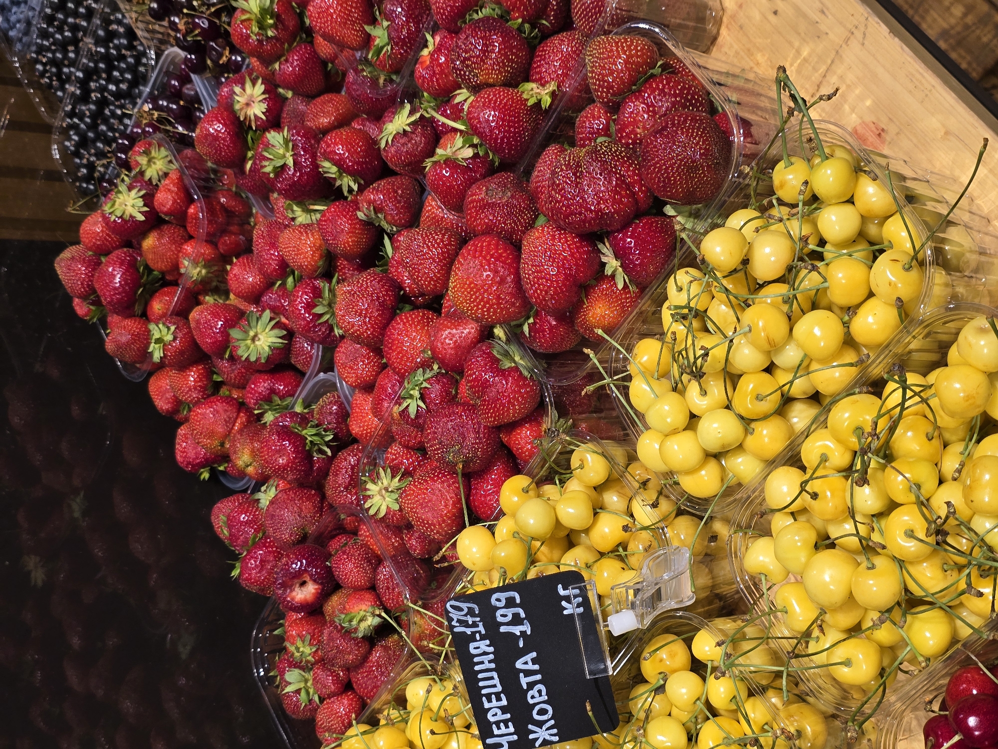 The image displays a vibrant overhead view of fresh fruit, primarily strawberries and yellow cherries, arranged in clear plastic containers. The strawberries are a rich red, some with visible green stems and leaves, filling several containers in the foreground and mid-ground. To the right and slightly further back, piles of bright yellow cherries are stacked in similar containers, their stems still attached. In the upper left corner, a container of dark, small berries, likely blueberries or currants, is partially visible. The fruits are laid out on what appears to be a wooden surface, suggesting a market stall or a retail display.
A small black price tag is affixed to one of the containers of yellow cherries, featuring white handwritten text in Cyrillic. The visible text reads "ЧЕРЕШНЯ - 179" and "ЖОВТА - 199 КГ". This translates to "Cherry - 179" and "Yellow - 199 KG". The lighting suggests it could be daytime, with the light casting slight shadows, indicating an indoor or shaded outdoor setting. There are no people visible in the frame. The overall impression is one of fresh produce ready for sale, likely at a farmer's market or grocery store in Petropavlivska Borshchahivka, Ukraine. The scene evokes a sense of freshness and abundance.