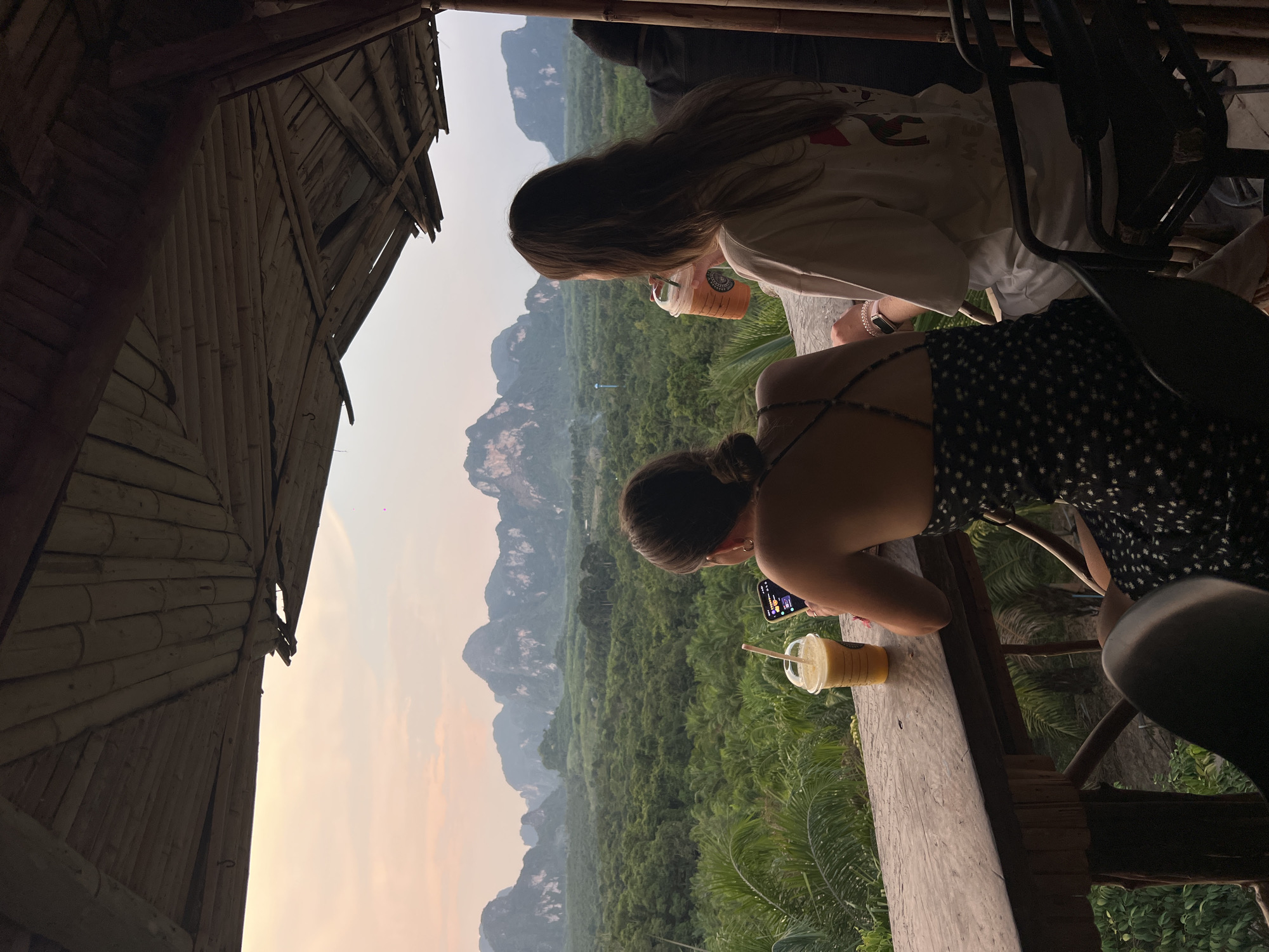 Two young women are at a high-altitude restaurant overlooking a green, mountainous landscape. The location is Khao Thong, Thailand. One woman with long brown hair is drinking from a tall, orange beverage cup with a straw. The second woman, with her hair in a bun, is looking at her phone and has a yellow beverage in a cup with a straw on the wooden railing. The restaurant structure is made of bamboo. A chair is visible in the lower right. The sky is light orange.