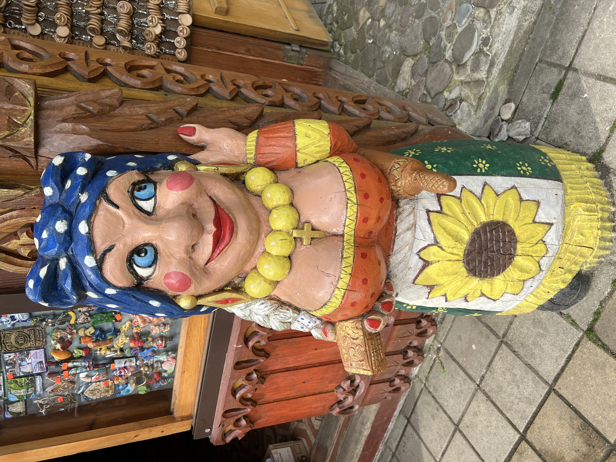 A painted wooden sculpture of a woman stands outside a building in Goryachiy Klyuch, Russia. The sculpture, depicting a woman with blue eyes, rosy cheeks, and a polka-dot blue headscarf, is situated on a shelf and is surrounded by souvenir trinkets inside a store. She is dressed in an orange top and a green and white skirt adorned with a sunflower. She wears a yellow necklace and holds a small box in her right hand. The background includes a stone wall, a wooden shelf with carved ornamentation, and a display of wooden keychains. The ground near the sculpture is paved with stone tiles and concrete.
