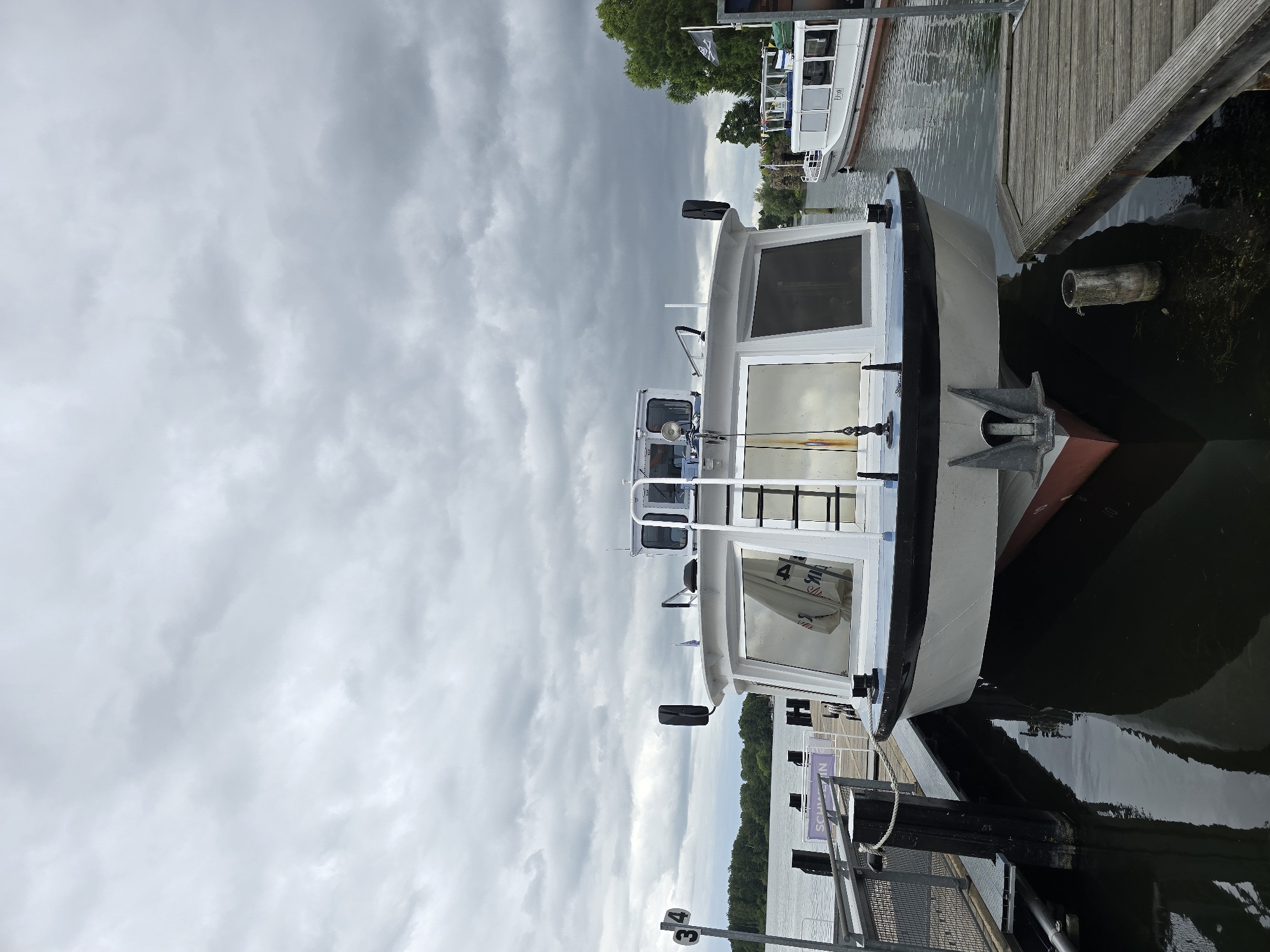 The image shows a white boat docked at a wooden pier on a cloudy day. The boat has large windows and a ladder leading to the upper deck. In the background, another boat and trees are visible. The scene appears to be in the city of Schwerin, Germany, as indicated by a sign that reads "SCHWERIN". The water is calm and reflects the boat and the sky. The overall atmosphere is peaceful and serene.