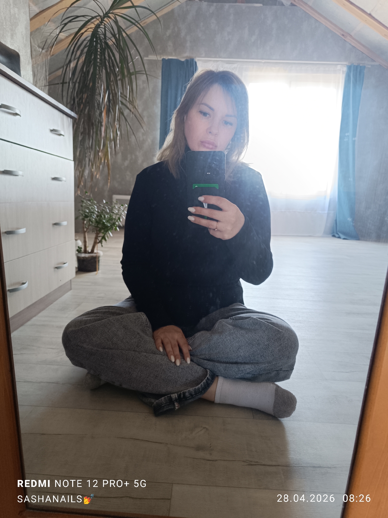 The image is a mirror selfie depicting a woman sitting cross-legged on a light wooden floor in an indoor setting, likely an attic room in Chaikovsky, Russia. She is holding a black smartphone with a green-accented case to capture her reflection. The woman has medium-length light brown hair and a neutral expression. She is dressed in a black long-sleeved top, loose grey pants, and white socks with grey toes. A small ring is visible on a finger of her right hand, which holds the phone.
The room features a sloping ceiling with exposed wooden beams and textured grey walls. To the left, a light-colored wooden dresser with multiple drawers and silver handles is partially visible. Two potted plants add greenery to the scene: a tall, leafy plant stands behind the dresser, and a smaller, bushier plant is positioned further back near the wall. A large window with light blue curtains dominates the background, allowing bright natural light to flood the room, causing some glare in the mirror. A radiator or vent is visible on the far wall beneath the window.
Visible text on the image includes the phone model "REDMI NOTE 12 PRO+ 5G" and the handle "SASHANAILS💅" in the bottom left corner. A timestamp "28.04.2026 08:26" is displayed in the bottom right corner, indicating the date and time the photograph was taken. The overall scene conveys a casual, brightly lit morning atmosphere.