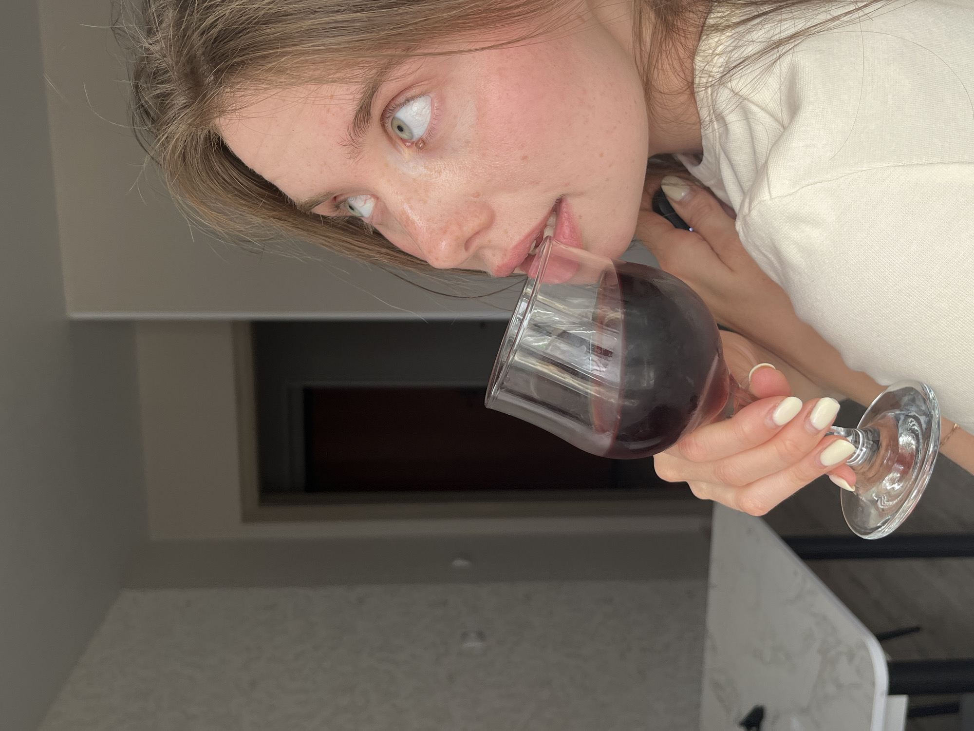 A young woman with fair skin and light brown hair is depicted in an interior setting. She is drinking red wine from a clear glass, looking upwards with wide eyes. She is wearing a light-colored shirt. The background appears to be a kitchen or dining area, with neutral-toned walls and countertops. The location is not identifiable as Chaikovsky, Russia from the visual information provided.