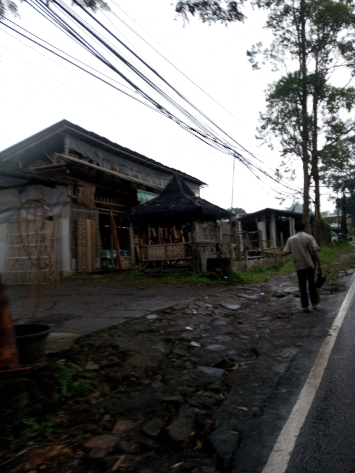 This image depicts an outdoor scene in the city of YonPhoogla, Bhutan, under an overcast sky. A single individual, viewed from behind, walks from the mid-ground towards the lower-right, along the shoulder of a dark, damp asphalt road that features a solid white line on its right edge. This person wears a light-colored, short-sleeved shirt and dark trousers, holding a dark object in their left hand. To the left of the road, an uneven, unpaved area of dirt, stones, and sparse green foliage separates the road from a series of structures.
In the mid-ground on the left, a two-story building with a dark, pitched roof and a facade constructed from wood and bamboo materials is visible. Its front exhibits open sections displaying what appear to be stacked wooden panels. Adjacent and to the right of this main building stands a smaller, open-sided structure featuring a distinctive dark, pointed thatched roof, with interior elements resembling bamboo railings and seating. Further right, another smaller, possibly incomplete, structure is partially visible behind a low wall.
Numerous dark utility lines crisscross horizontally across the upper portion of the frame against a bright, uniformly overcast sky. On the far right side of the road, tall, slender trees with green foliage rise into the upper frame. In the lower-left foreground, a dark-colored bucket or barrel is positioned next to a reddish-brown cylindrical object. The ground surface throughout the scene appears damp.