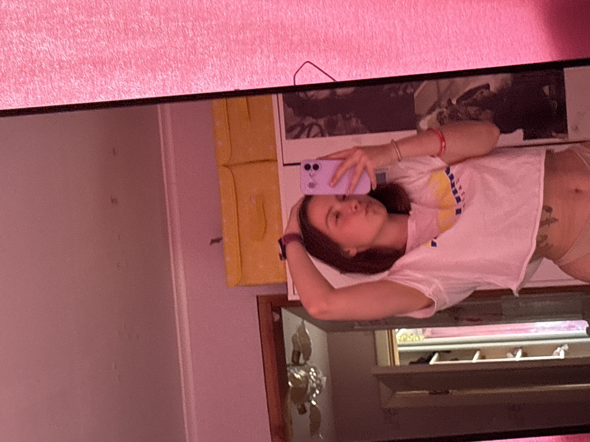 A young woman is taking a selfie in a mirror. She is wearing a white t-shirt and holding a purple phone. The room appears to have pink walls, and there is a yellow object on the wall behind her. The location appears to be in a domestic interior, as the presence of a closet suggests, but I cannot confirm the city of Прикамский, Russia.