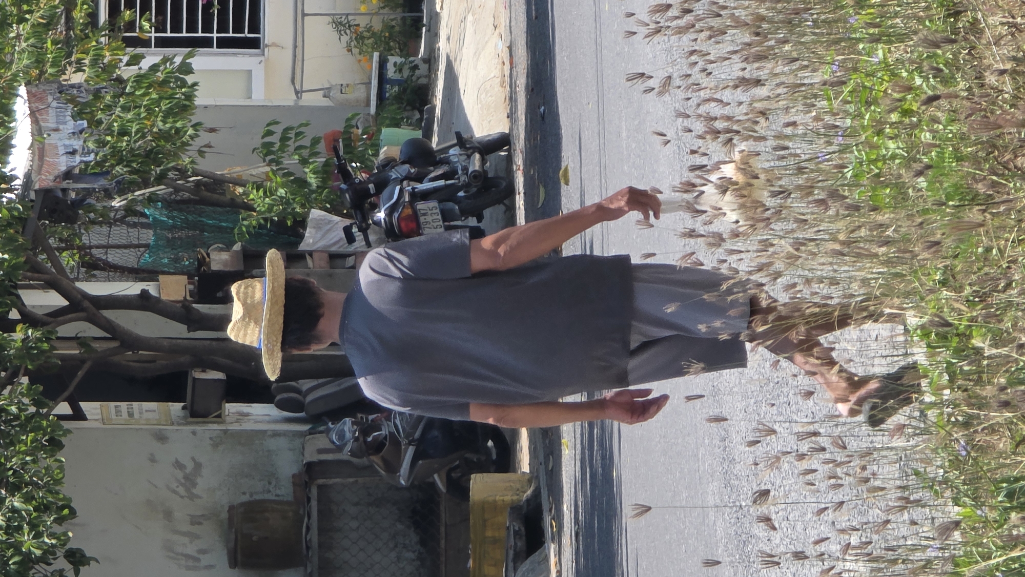 A man wearing a straw hat, gray shirt, and gray shorts is walking barefoot on a paved street. To his left are buildings and a motorcycle. To his right, weeds and grass. The motorcycle's license plate says "79-N7 5035". The photo was taken in Bắc Nha Trang, Vietnam.