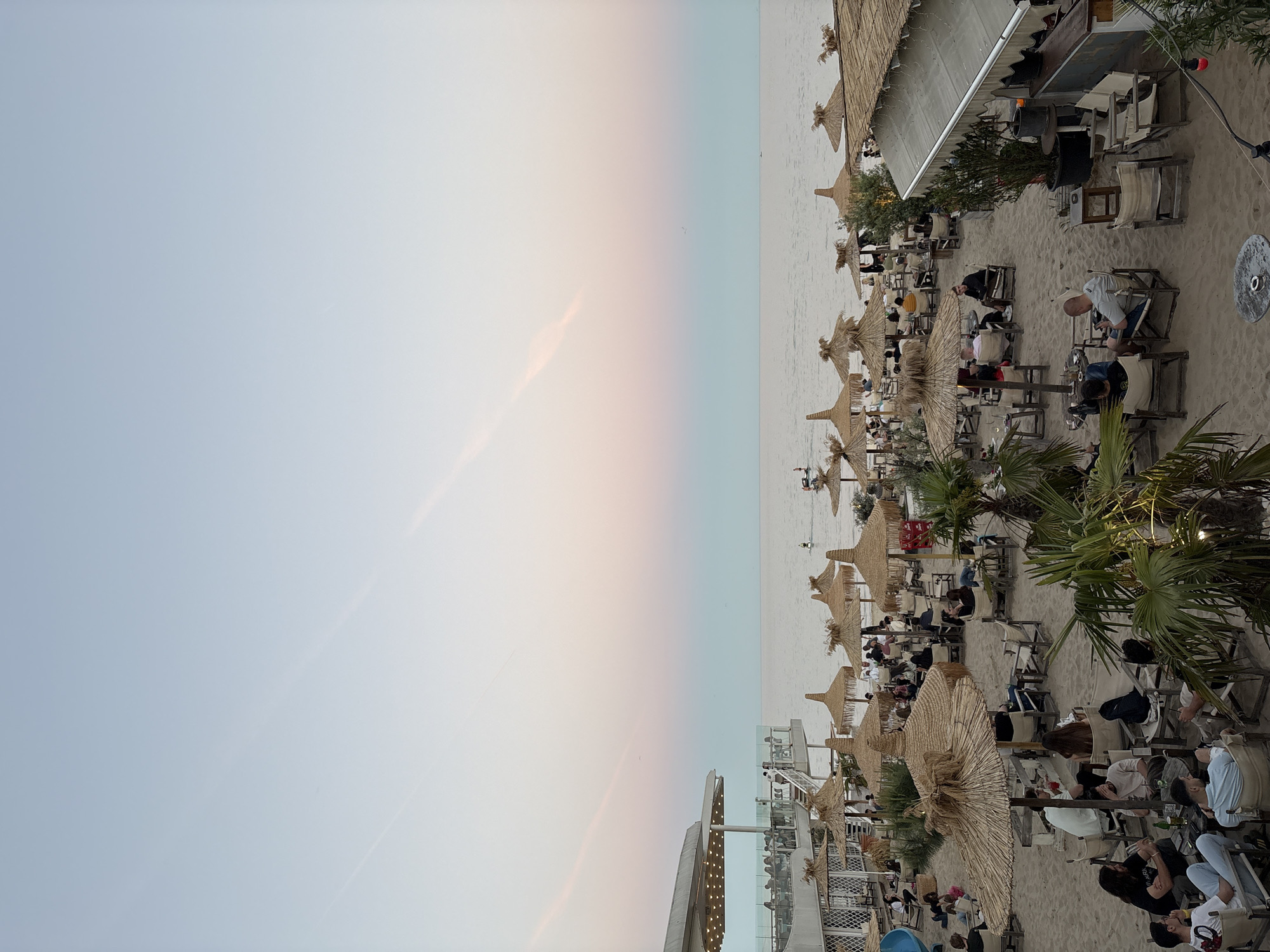 The image shows a beach scene in Varna, Bulgaria. People are seated at tables and chairs under straw umbrellas on a sandy beach. There are palm trees and a multi-story structure with lighting. The sky is light blue to pink near the horizon, suggesting it is either sunrise or sunset. The sea is calm.