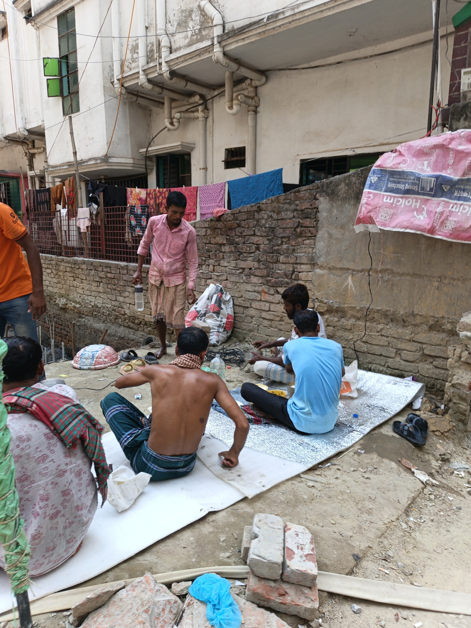 In Dhaka, Bangladesh, several men are gathered outdoors on a plot of sandy ground next to a partially constructed building. One man, shirtless and wearing a patterned sarong, sits cross-legged with his back to the camera. Another man in a blue t-shirt also sits cross-legged on a reflective mat, eating. A third man in a pink shirt stands holding a water bottle. Other men are also present, some partially visible in the background. Clothing lines hang from the upper floors of the building, and a tattered banner with the word "Holcim" is affixed to the side of the building. Bricks and other debris are scattered around the ground.