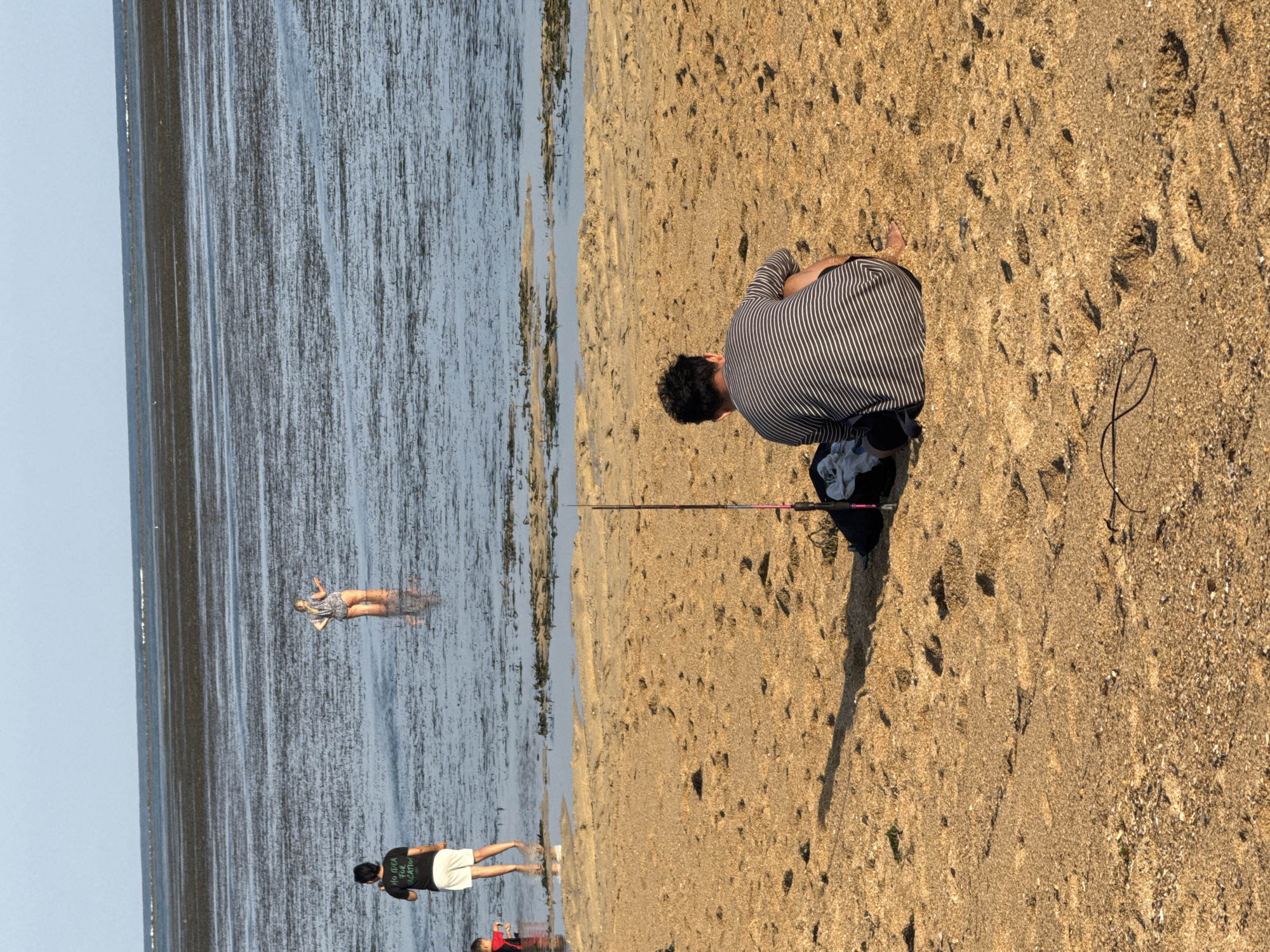 The image captures a beach scene in Denpasar, Indonesia, likely at low tide given the expanse of shallow water and exposed seabed. In the foreground, a person is sitting on the sand facing away from the camera, dressed in a striped t-shirt and shorts. Next to them, a fishing rod is propped up, leaning against a bag. The sand is covered in footprints. In the mid-ground, a man in a black t-shirt and white shorts stands in the shallow water, facing towards the sea. To his left, a child in a red garment is partially visible wading. Further out, a person in a light-colored patterned outfit stands in the water, also facing away. The background consists of a calm sea with a distant horizon.