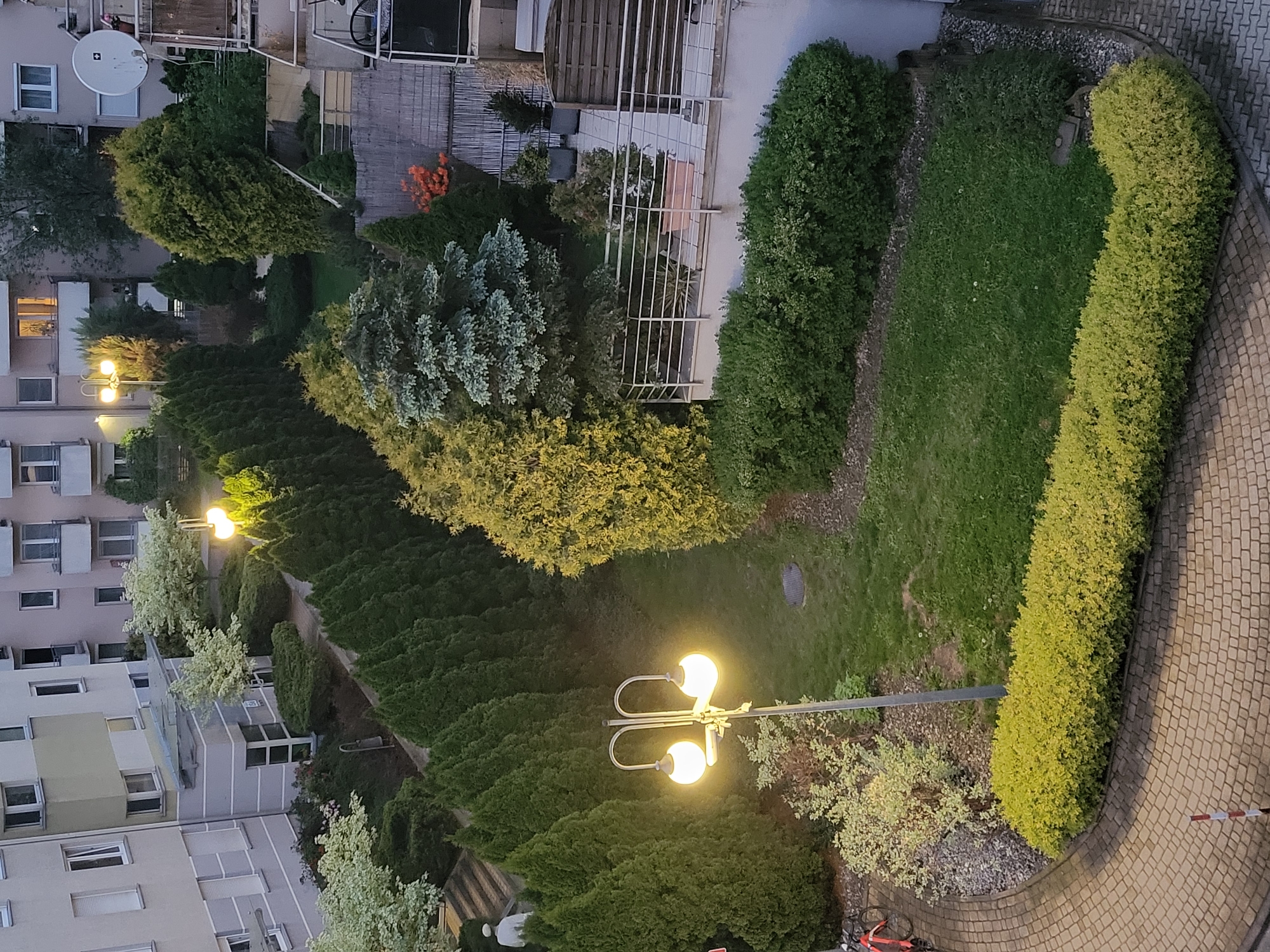 The image depicts an aerial view of a residential area in an unknown city. The scene includes apartment buildings on the left side of the image, interspersed with greenery. A row of coniferous trees extends from the bottom to the middle of the frame, illuminated by street lamps. A paved path curves along the right side of the image, lined with bushes and vegetation. A green lawn and more trees occupy the central space. The scene is captured during twilight, with streetlights providing illumination.
