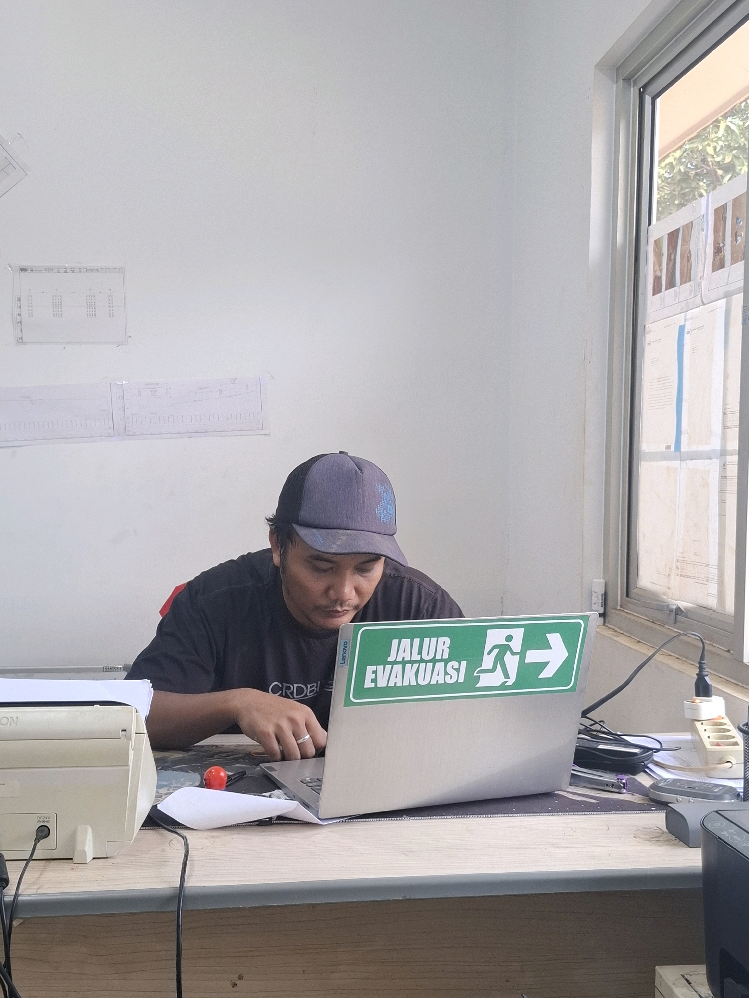 In a brightly lit room with white walls, a person wearing a dark t-shirt and a grey baseball cap is seated at a wooden desk, engrossed in using a silver laptop. The laptop displays a green sticker with white text and an icon of a person running towards an arrow, which reads "JALUR EVAKUASI". To the left of the laptop is a white printer with cables plugged into it. A red object, possibly a pen or a small toy, rests on the desk. Various papers and office supplies are scattered across the desk and on shelves on the wall behind the person. A window on the right side of the frame reveals a glimpse of green foliage and a cloudy sky. The overall setting appears to be an office environment, possibly in Ciheulang Tonggoh, Indonesia.