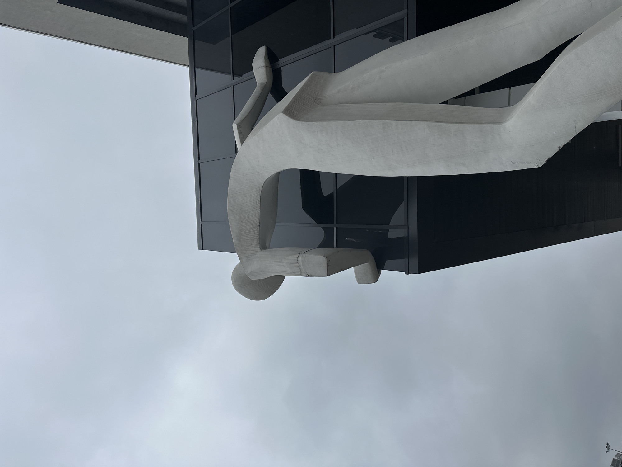 The image shows a sculpture of a stylized human figure mounted on the facade of a modern building with dark, reflective glass windows in Nizhny Novgorod, Russia. The sculpture is made of a light-colored material, possibly concrete or stone, and depicts a human form with simplified, rounded features. The figure appears to be seated or suspended. The sky is overcast and gray. The building is modern in style, with clean lines and a dark facade. The sculpture is positioned prominently on the building's exterior.