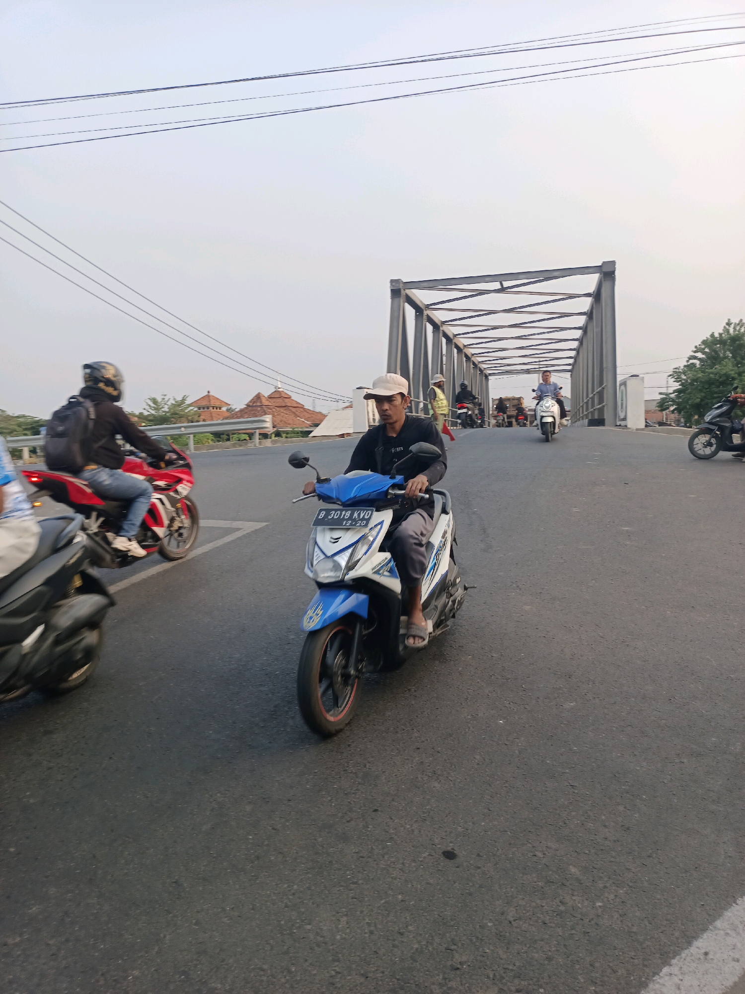 The image captures a busy street scene on a bridge in Karawang, Indonesia, with several motorcycles navigating the roadway. In the foreground, a man wearing a light-colored cap and a dark t-shirt rides a blue and white scooter. To his left, another person on a red motorcycle with a black backpack is partially visible. Further down the bridge, other motorcycles and vehicles are seen, indicating moderate traffic. The bridge itself has a distinct metal truss structure overhead. The background features buildings with reddish-brown tiled roofs and lush green trees, suggesting a semi-urban or suburban setting. The sky is overcast with a soft, diffused light. The overall atmosphere is one of daily commute and activity on a slightly hazy day.