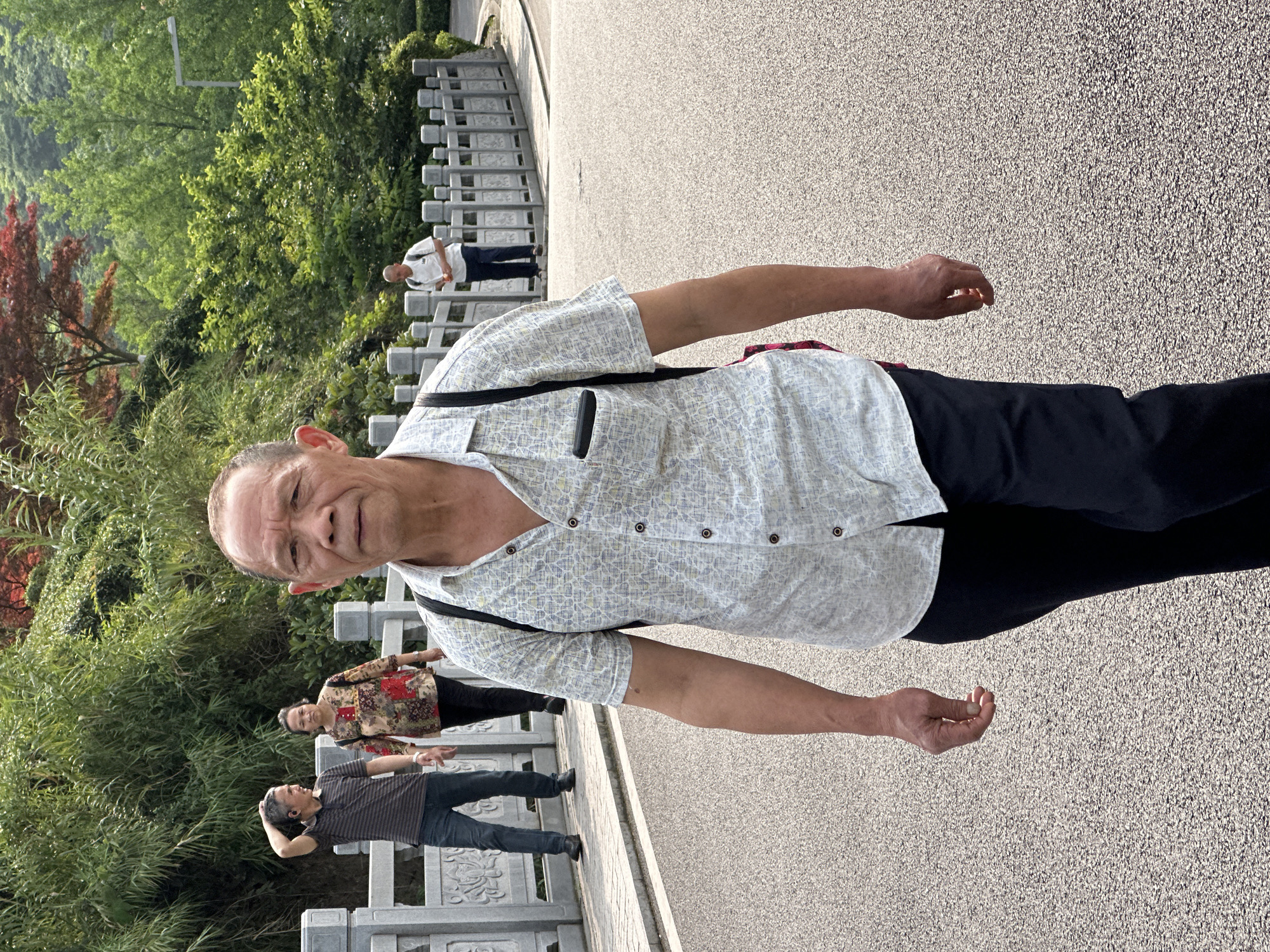 A man wearing a patterned short-sleeved shirt and dark trousers stands on a grey concrete surface with what looks like a bridge railing to his left. Behind him, other people are on the bridge. There is lush green vegetation, and trees are in the background. Based on the landscape and architecture, this may be a park area within Liangjiang, China.